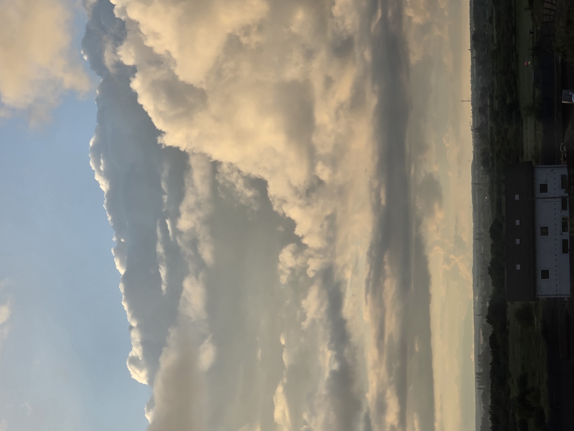 A wide-angle shot captures a dramatic sky filled with large, fluffy clouds illuminated by the setting sun. The clouds range in color from bright white and yellow on the right side of the frame to muted grays and blues on the left. Below the expansive sky, a flat landscape stretches across the horizon, featuring dark green trees and fields. In the foreground, a white building with a dark roof and multiple windows is partially visible on the right. The building appears to be under construction or unfinished. The distant horizon is hazy, with faint outlines of structures and trees. The scene suggests a rural or semi-rural area, possibly on the outskirts of a city.