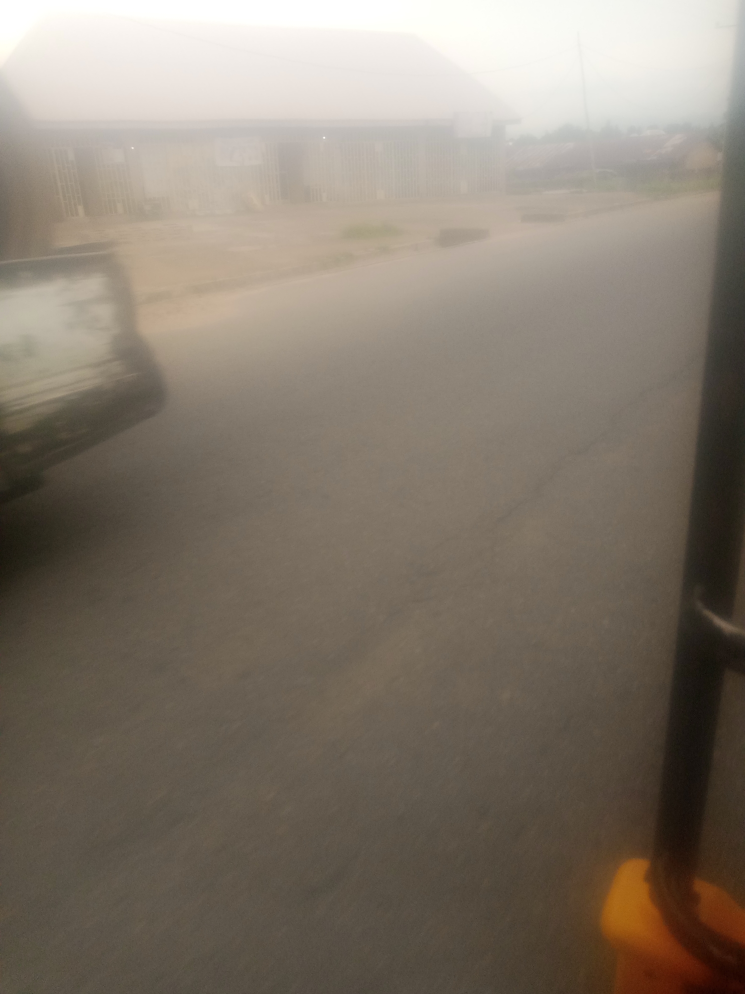 The image shows a slightly foggy street scene in Jalingo, Nigeria. On the left, a part of a vehicle, possibly a truck, is visible. Across the street, there is a building with a row of windows and vertical bars, suggesting it might be a shop or office. The road surface is asphalt. In the foreground on the right, part of a dark vertical pole and a yellow object, possibly a container or equipment, are visible. The sky is hazy and bright, indicating daylight. The overall scene appears to be a typical street in a Nigerian city with some commercial activity.