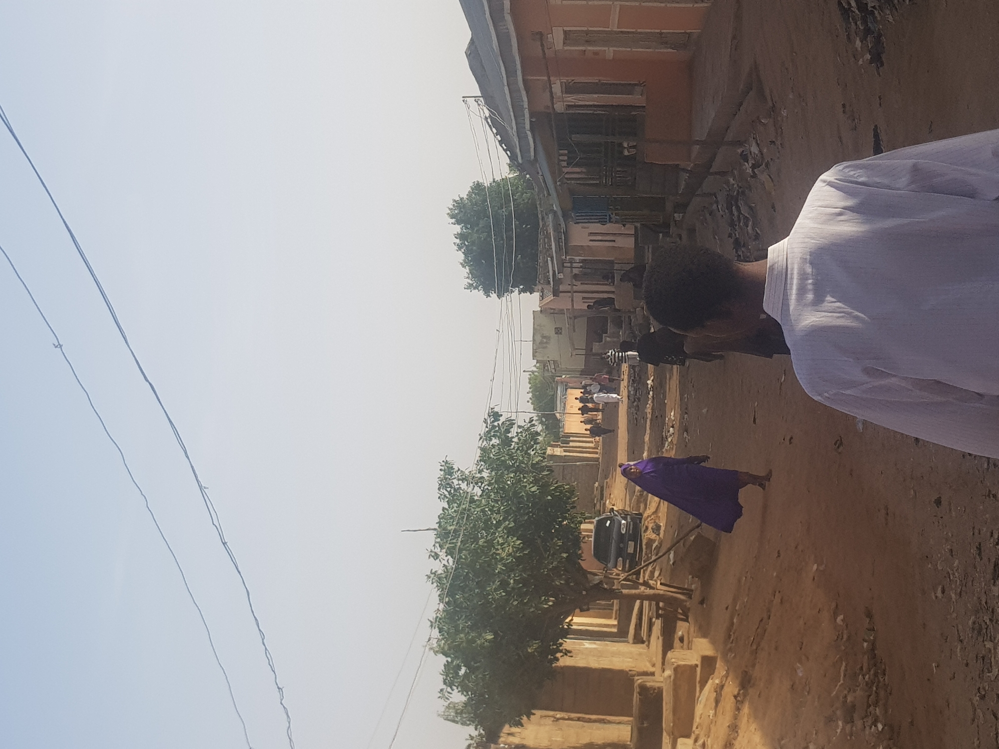 The image depicts a street scene in Dutsinma B, Nigeria. The unpaved street is lined with buildings of varying heights and materials. A person in a white shirt is partially visible in the foreground on the right. Further down the street, a woman in a purple robe is walking. Other individuals are seen in the distance. A car is parked near a tree along the street. Overhead power lines are visible against a hazy sky.