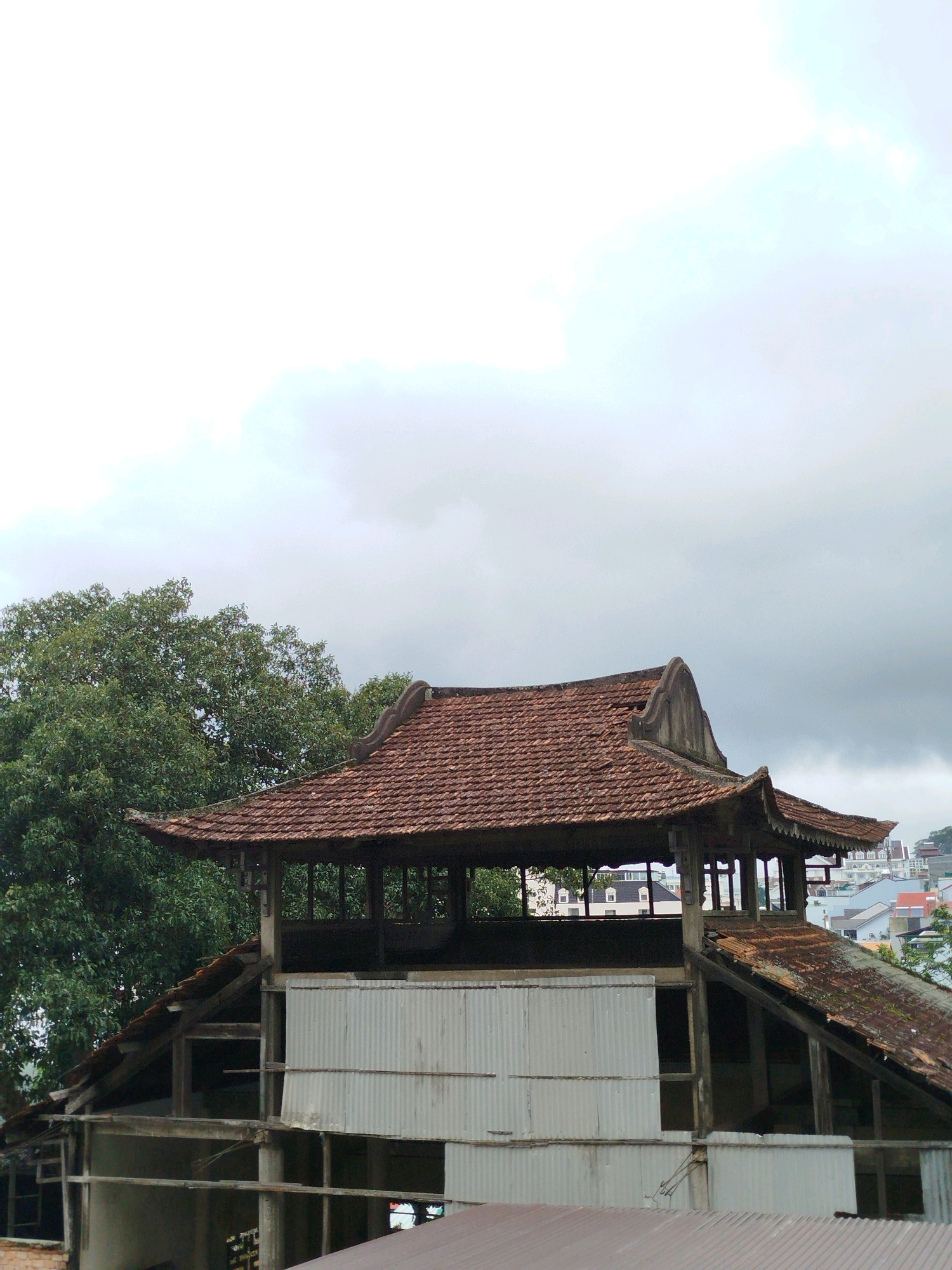 The image shows an unfinished building with a traditional tiled roof structure in Da Lat, Vietnam. The main focus is on the upper story, which features a multi-tiered, curved roof covered in reddish-brown tiles. The roof is supported by wooden pillars and has decorative eaves. Below this tiled structure, the building appears to be in a state of construction or disrepair, with exposed wooden beams and corrugated metal sheeting forming some of the walls. A large, leafy green tree is visible to the left of the building. In the background, other buildings and structures of the city can be seen, suggesting an urban or semi-urban setting. The sky is overcast with gray clouds.