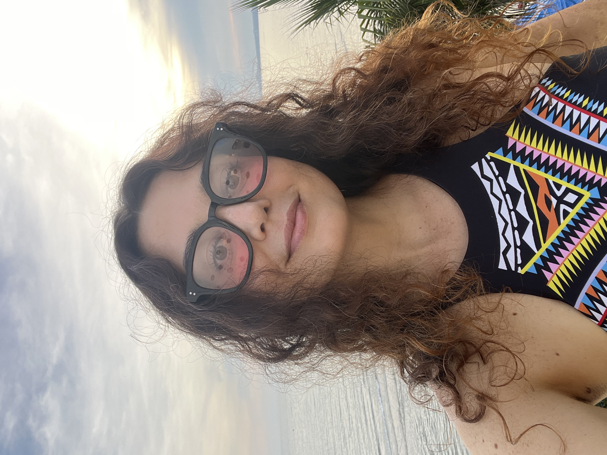 A woman with long, wavy brown hair and fair skin is pictured in a close-up shot, wearing black-rimmed glasses and a black swimsuit with a colorful geometric pattern. Her gaze is directed towards the camera, and she has a slight smile. The background shows a body of water with gentle ripples, possibly the ocean, under a sky with soft clouds and warm lighting, suggesting it might be late afternoon or early morning. A hint of palm fronds is visible at the top of the frame, indicating a tropical or coastal setting. The image has been rotated 90 degrees counter-clockwise. No visible text is present.
