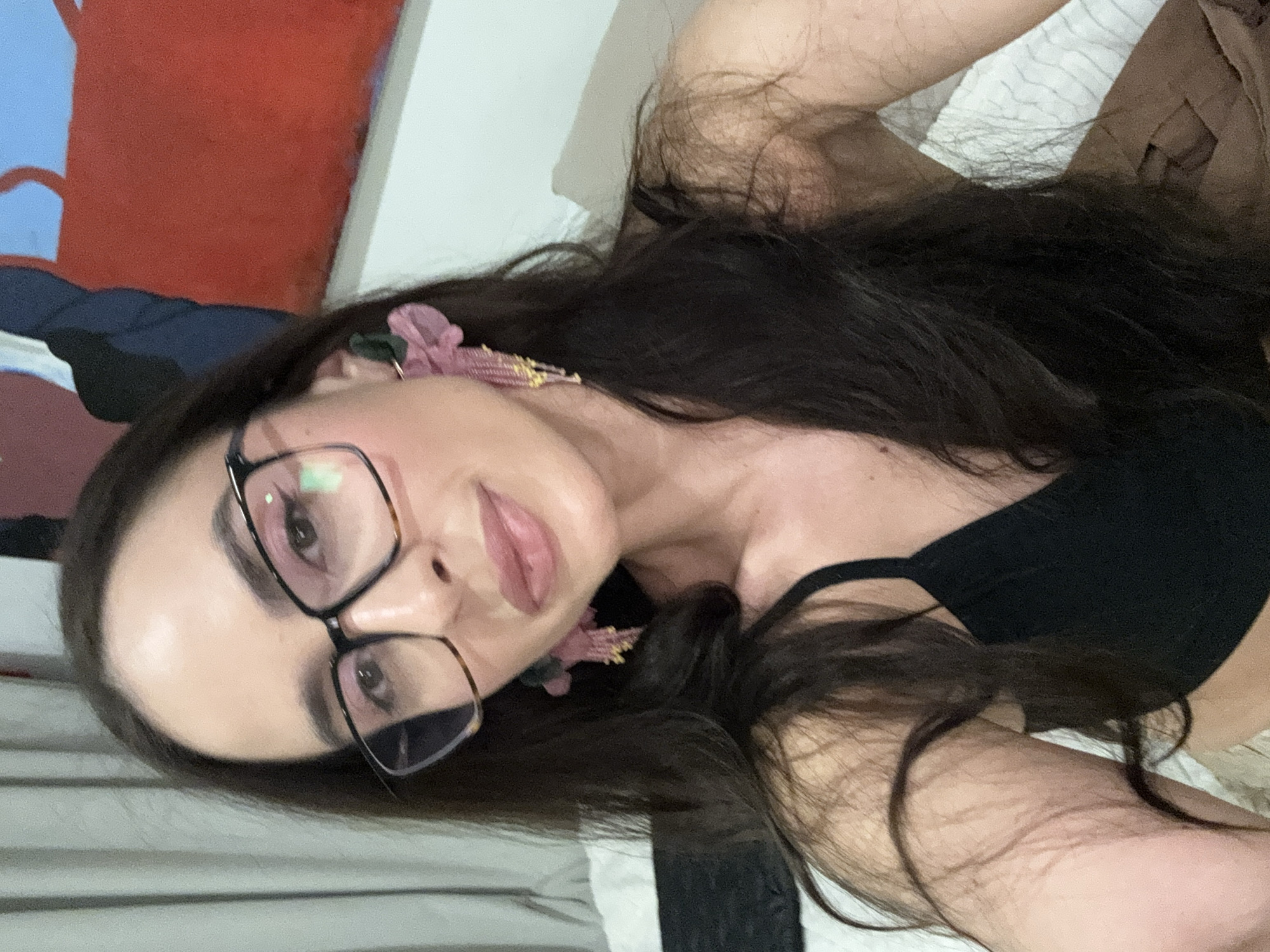 A young woman with long, dark hair is captured in a selfie indoors, located in Krasnoyarsk, Russia. She wears black-rimmed glasses and a black top or bra. Notable accessories include large, pink or purple ornamental earrings, possibly with beaded or tassel details. Her lips are slightly parted, conveying a neutral to subtly alluring expression as she looks directly at the camera. The background features a bed with striped and brown bedding, and a wall adorned with a red and blue abstract painting or wall art. The lighting suggests an indoor setting; the specific time of day and external weather are not discernible. No text is visible in the image.
