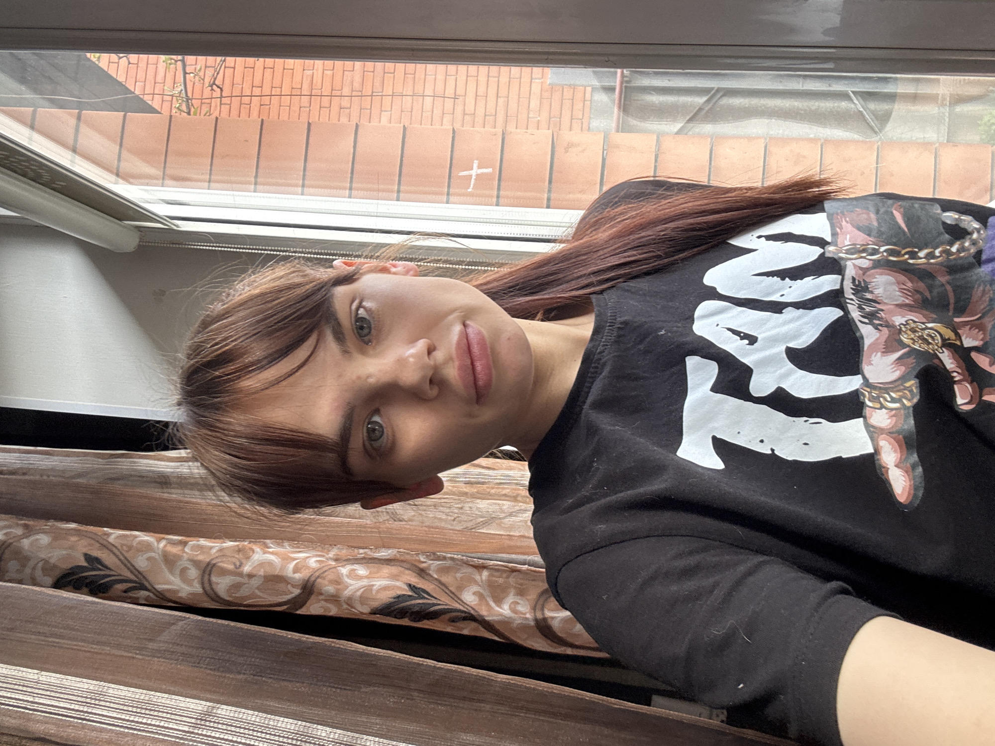 A selfie-style image features a fair-skinned female with light brown hair and a fringe posing indoors near a window. Her hair is parted to the side and cascades towards her right shoulder. She wears a black t-shirt with a prominent white graphic design, and a stylized image involving a ring and chain.
Behind her, a window reveals a brick wall and vegetation outside. The interior includes window blinds with a patterned design. The environment suggests an apartment setting. There is no information to confirm the location is Rostov-on-Don, Russia.