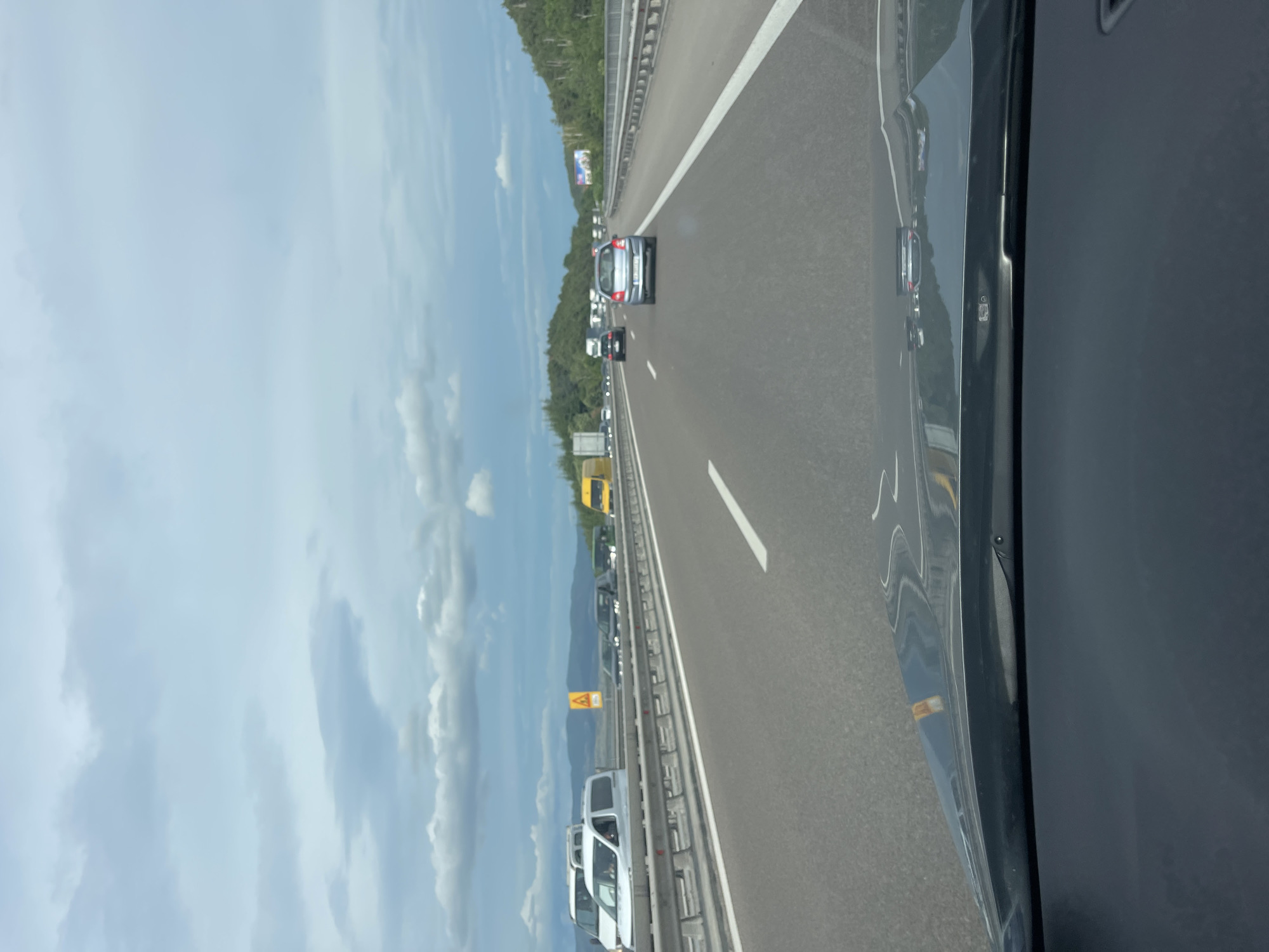 The image depicts a view from the driver's perspective of a highway. The road is paved with asphalt and has white lane markings. There is traffic on the highway, with several cars and vans visible in front and in other lanes. The vehicles include a silver hatchback, a yellow van, and several white vans. In the distance, there are trees and hills, suggesting a rural or suburban setting outside of a city. The sky is cloudy with a mix of grey and white clouds. There is a metal guardrail on the side of the highway. A yellow triangular sign with a black symbol is visible on the left side of the road. The dashboard of the car is visible in the foreground, reflecting the highway and sky. The scene is consistent with a highway in or near Gorno Varshilo, Bulgaria, given its general appearance of a European highway with surrounding nature.