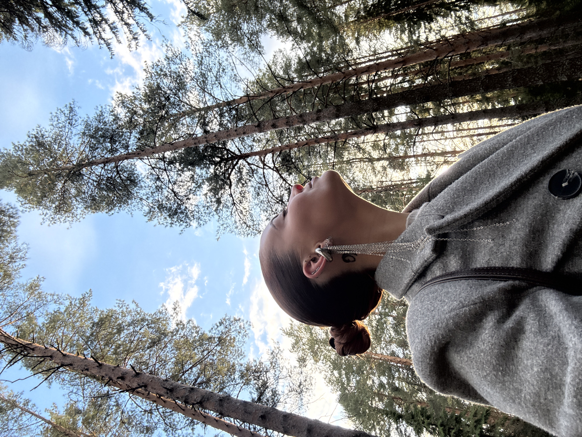 This low-angle photograph, taken in Прикамский, Russia, captures a person looking upwards into a pine forest on a bright, clear day. The main subject, positioned on the right side of the frame, is a person (likely female) seen from the neck up in profile. Their dark hair is styled in a neat bun, and they wear an intricate silver ear cuff with long, delicate chains hanging down, along with a small, circular tattoo below their ear. Red lipstick is subtly visible. They are clad in a grey, textured coat or jacket featuring a dark, circular button.
The background is dominated by the tall, straight trunks and dense green needles of pine trees, extending high into a clear, vibrant blue sky dotted with soft white clouds. Sunlight filters through the canopy, creating a dappled effect. The perspective is from directly below, emphasizing the height of the trees and the expansive sky. The person appears to be in a moment of quiet contemplation or appreciation of the natural surroundings. No visible text is present. The time of day seems to be bright daylight, likely mid-day.