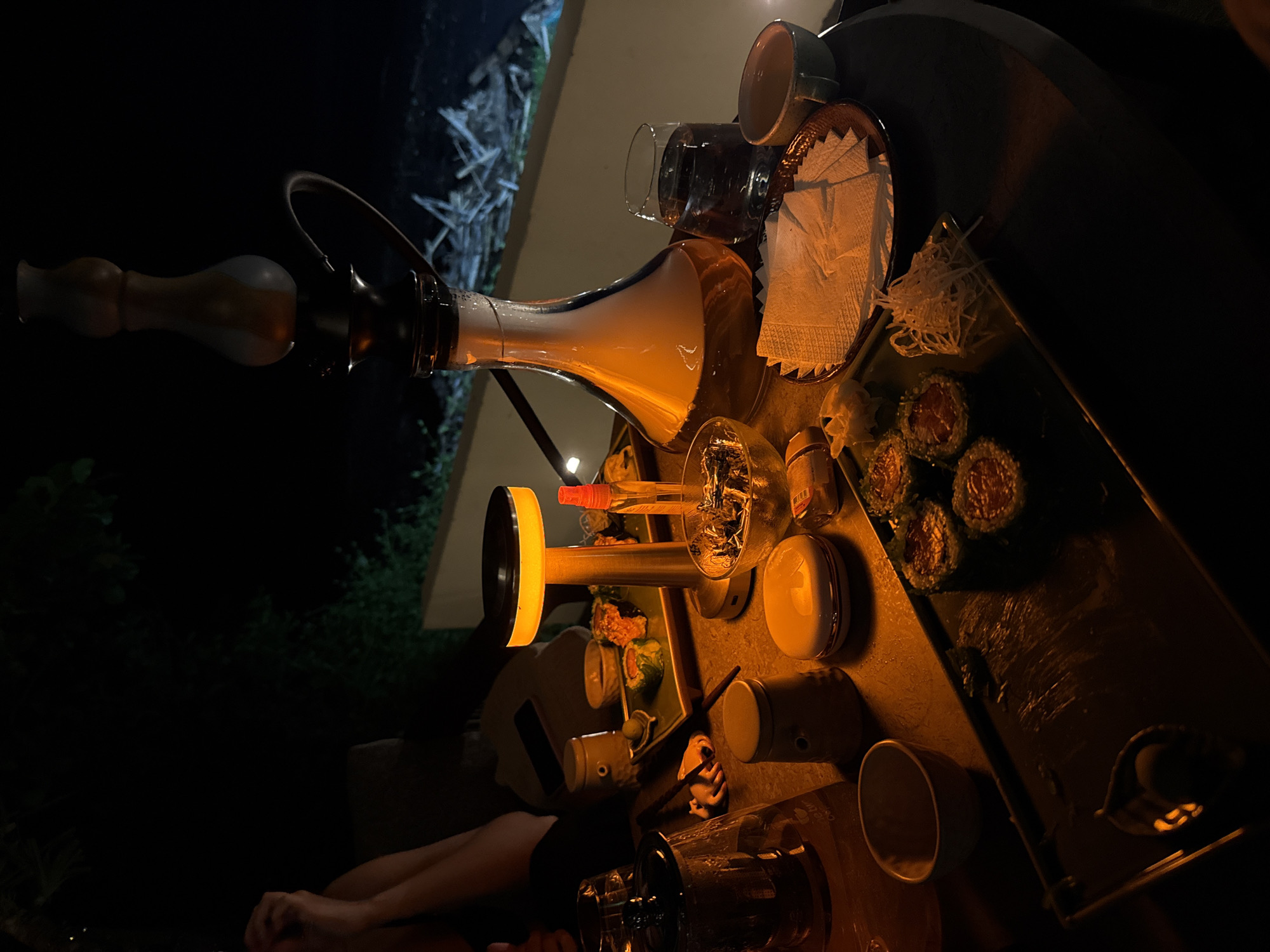 The image captures an outdoor night scene, likely a casual social gathering, focused on a table laden with food and drinks, illuminated by warm artificial light. At the center, a modern hookah with a transparent, glowing base and a dark stem is prominently displayed.
A dark table holds various items, bathed in the golden glow of a cylindrical lantern or light source. On the table, several pieces of sushi or maki rolls, accompanied by shredded daikon radish, are arranged on a rectangular plate. Beverages include a glass containing liquid, a tumbler, and several ceramic cups. An ashtray filled with cigarette butts and ashes is also visible, suggesting smoking is taking place. White napkins are neatly stacked on a small, decorative plate, alongside other small bottles and containers.
Part of a person's arms and torso are visible in the lower left quadrant, indicating someone is seated at the table. The background is dark and out of focus, showing indistinct foliage and possibly distant structures, confirming the outdoor, nocturnal setting. The warm lighting and the arrangement of items create a cozy and relaxed atmosphere for an evening leisure activity. No discernible text is visible within the image.