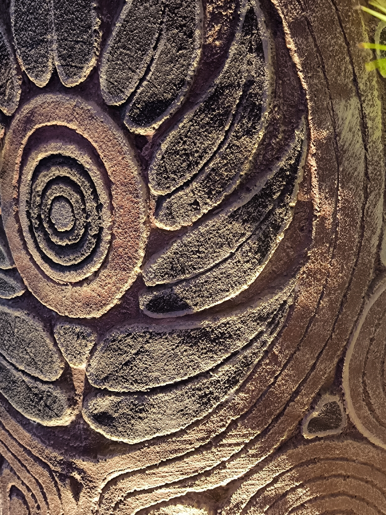 The image shows a close-up of a textured surface with an embossed pattern. The pattern consists of concentric circular shapes and curved, petal-like elements. The surface material appears to be stone or concrete, with a rough, granular texture. The lighting highlights the raised portions of the pattern, creating shadows that emphasize its three-dimensional quality. The colors are predominantly earthy tones of brown and gray, with some reddish hues in the circular elements. There is no indication of a specific location in Kediri, Indonesia, or any people, actions, or interactions within the frame. The image is purely a study of the abstract pattern and texture of the object.