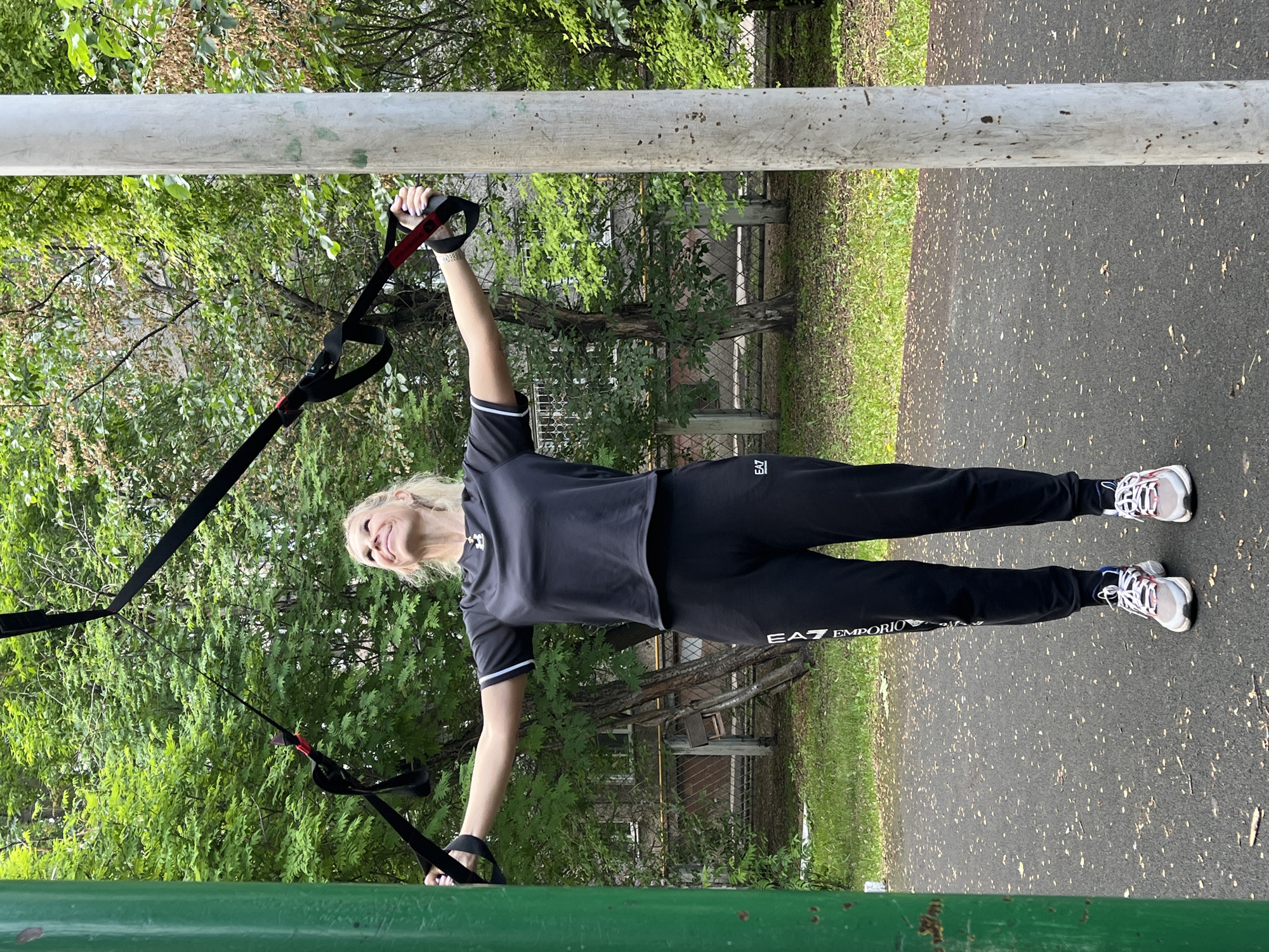 A middle-aged Caucasian woman with blonde hair is performing a TRX workout outdoors. She is wearing a black t-shirt, black track pants with white lettering, and athletic shoes. Her arms are extended to the sides, holding onto the TRX straps, which are anchored above. She is looking up with a smile. The workout appears to be taking place in an outdoor recreational area, possibly a park or schoolyard, with trees and grass in the background. A green pole is visible on the left and a grey pole on the right. The ground is paved with asphalt. The weather appears to be overcast.