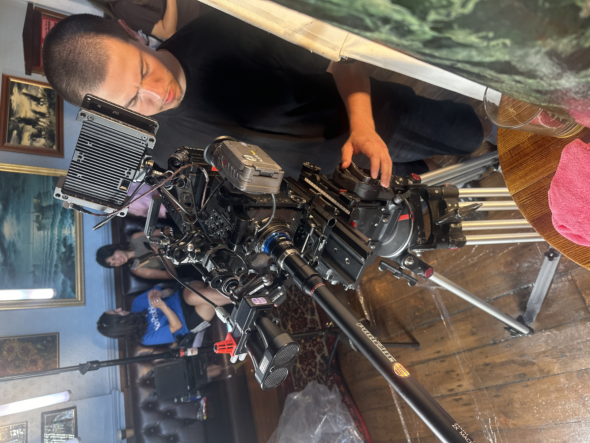 A cluttered interior scene shows a film crew preparing a shot. In the foreground, a professional film camera mounted on a black tripod dominates, being adjusted by a man in a black shirt. The camera is complex, with multiple components and cables attached. In the background, two women are seated in the room, one wearing a blue shirt with writing. The walls are adorned with framed pictures, and the floor is wooden. A light and other equipment are scattered around. Details of the exact location in London are indiscernible.