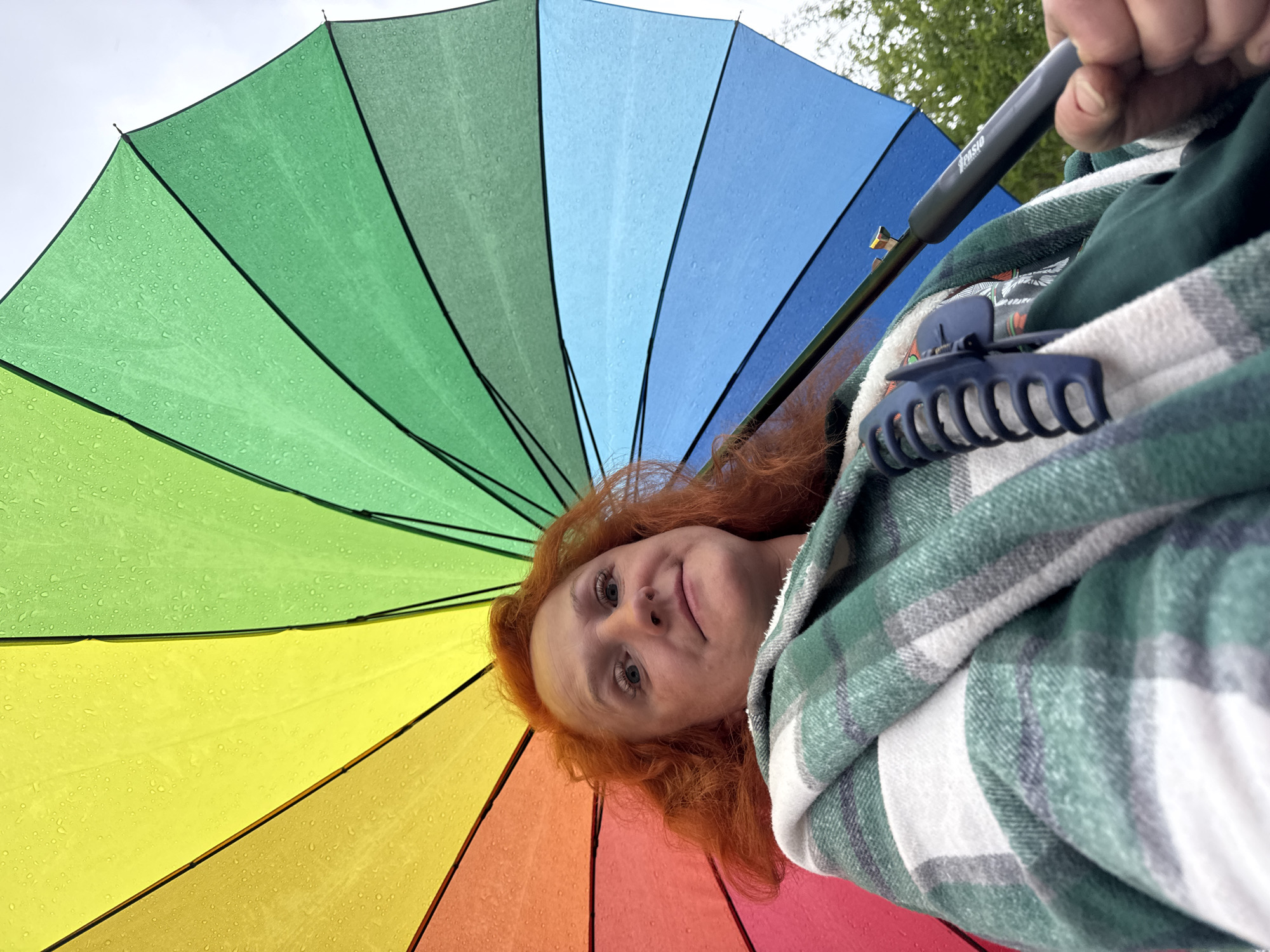 A woman with red hair is holding a rainbow umbrella. She is wearing a green and white plaid shirt. A dark blue hair clip is attached to the shirt. The background is out of focus, but appears to be trees. The photo was taken from a low angle, looking up at the woman and the umbrella. The location is unknown, and there is no confirmation that the photo was taken in Chaikovsky, Russia.
