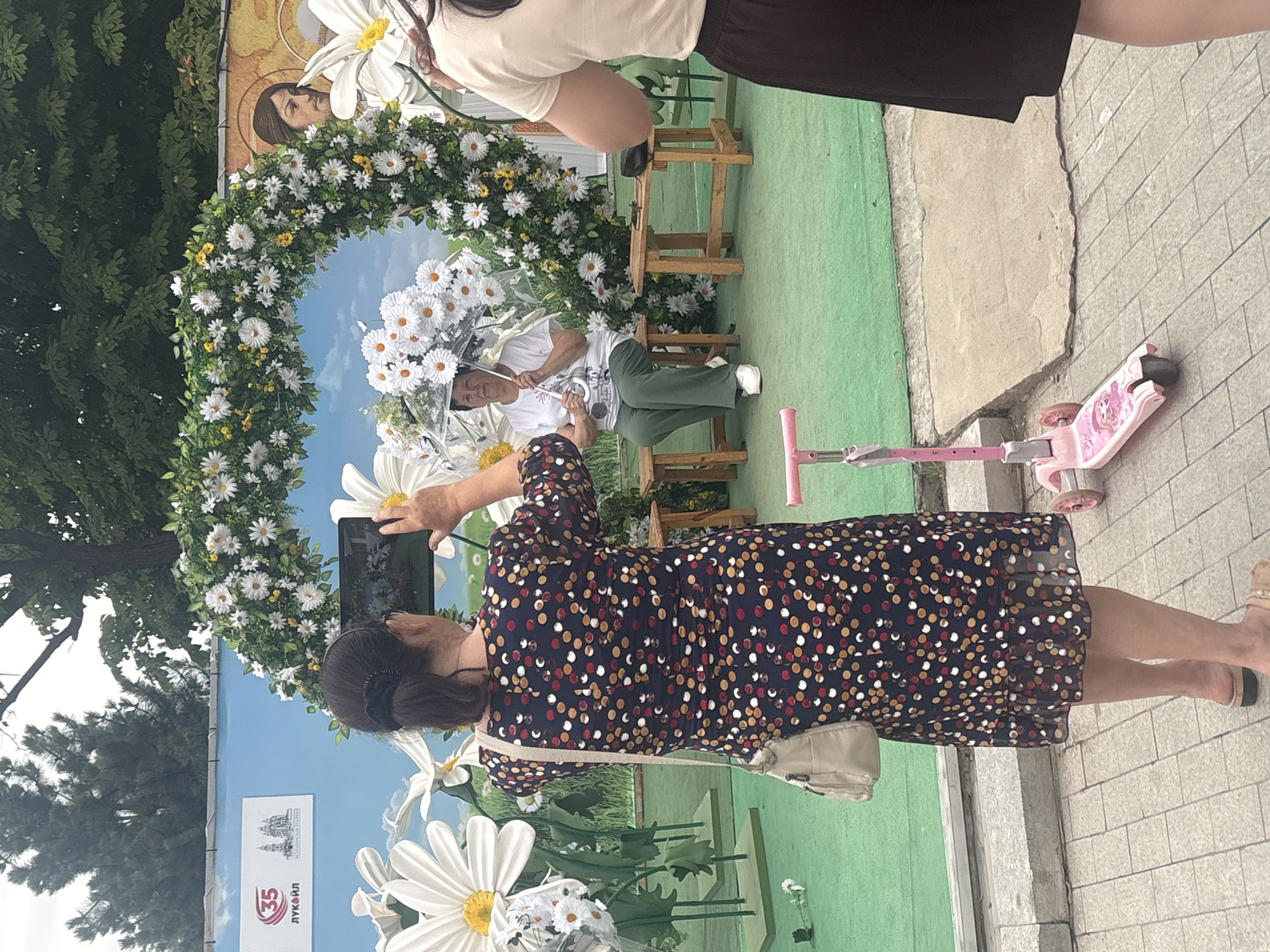 In Astrakhan, Russia, a woman in a dark dress patterned with colorful polka dots holds up a tablet to take a picture of a floral archway decorated with white daisies and greenery. The arch frames a backdrop that appears to be a mural of a blue sky with clouds and large white daisies. Inside the archway, a woman sits on a wooden bench, holding a clear umbrella adorned with white flowers. She is wearing a white t-shirt and green pants. To the right of the scene, a pink child's scooter rests on a tiled sidewalk. A person, partially visible in the upper right, wears a light-colored shirt and dark skirt. The overall impression is a pleasant, outdoor setting, possibly a park or public square, where people are enjoying a decorated space. The lighting suggests it is daytime. Visible text on a banner behind the floral arch includes "35" and "ЛУКОЙЛ", indicating a connection to the Lukoil company and possibly an anniversary or event related to the Astrakhan region.