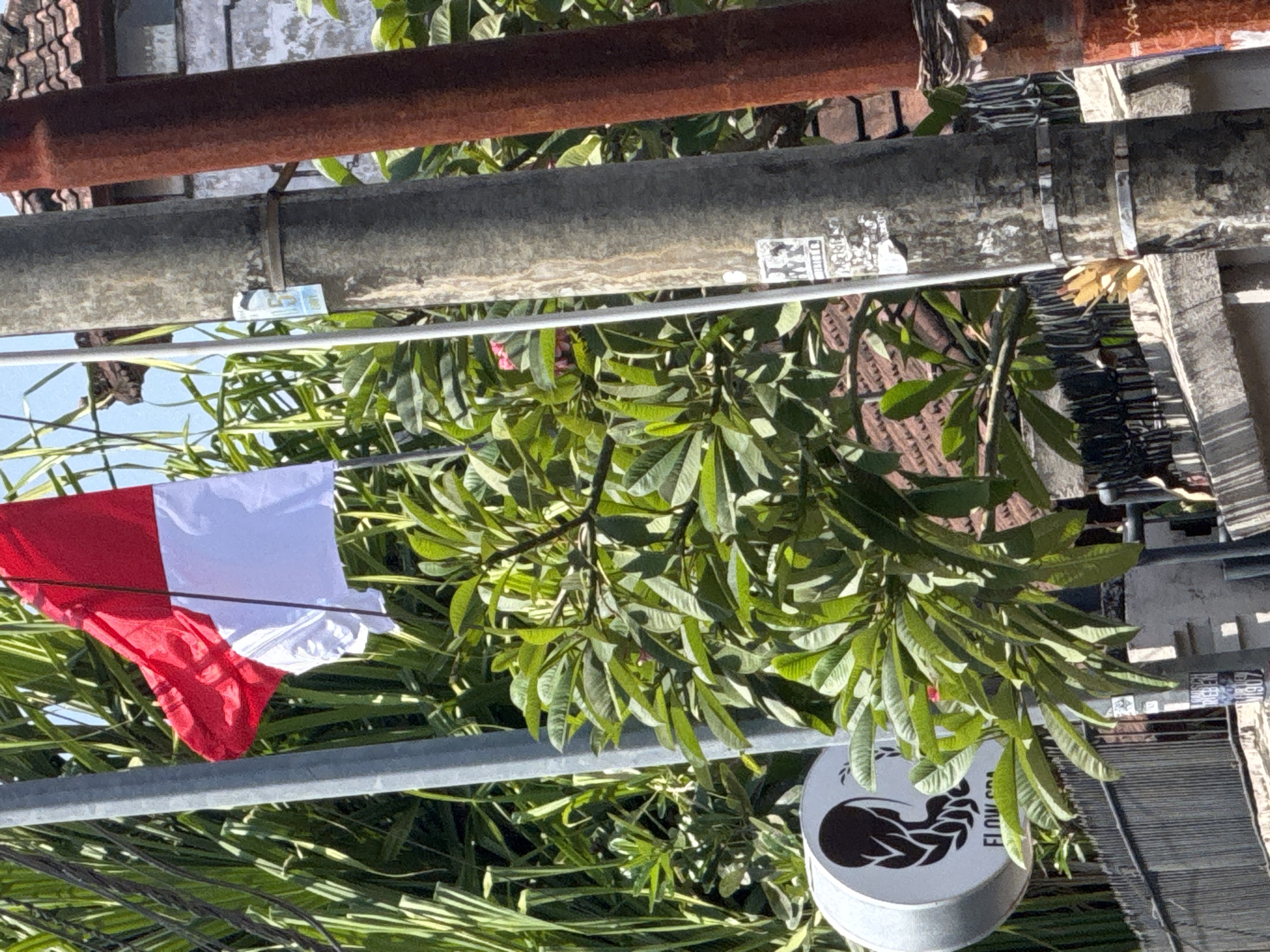 The image shows an Indonesian flag (red and white) waving against a backdrop of lush green foliage and a bright blue sky. The flag is attached to a pole which is partially obscured by other elements in the frame. To the right of the flag pole, a rusty brown pipe stands parallel. In the foreground, a gray utility pole is visible, adorned with several stickers. Below the flag and utility pole, a circular sign with a black silhouette of a head and shoulders, and the text "FLOW SPA" is mounted. The background reveals more dense greenery and a tiled roof structure. The overall scene suggests an outdoor environment in a tropical location, likely Kuta, Indonesia, given the presence of the Indonesian flag and tropical vegetation.