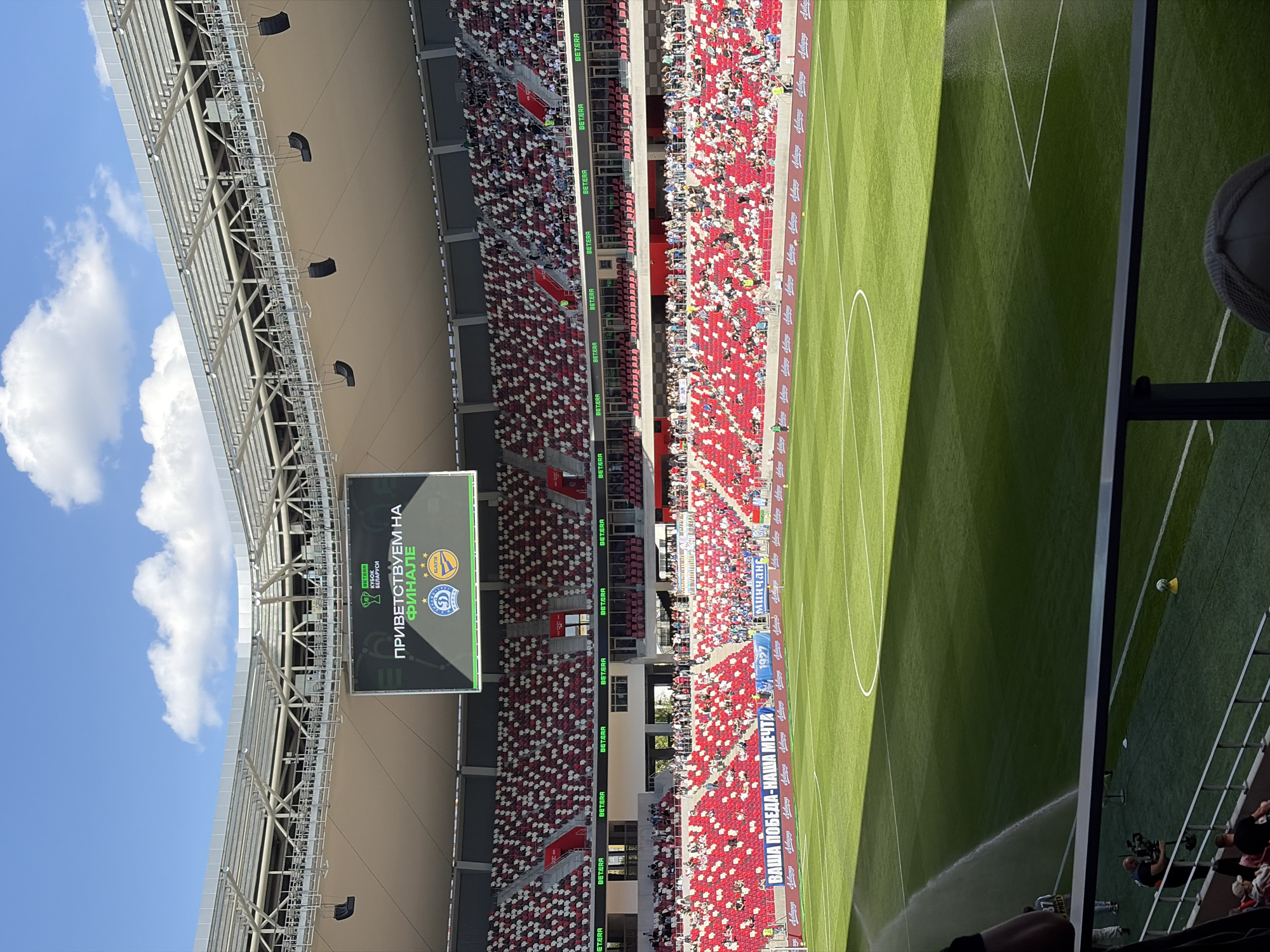 The image depicts a soccer stadium in Minsk, Belarus. The stadium is filled with spectators. A large scoreboard displays the text "ПРИВЕТСТВУЕМ НА ФИНАЛЕ" ("Welcome to the final"). The field is well-maintained and partially watered. Advertisements for "BETERA" and "Минчань" (Minsk) are visible around the stadium. The text "ВАША ПОБЕДА-НАША МЕЧТА" ("Your victory is our dream") is also displayed. A soccer ball is on the ground.