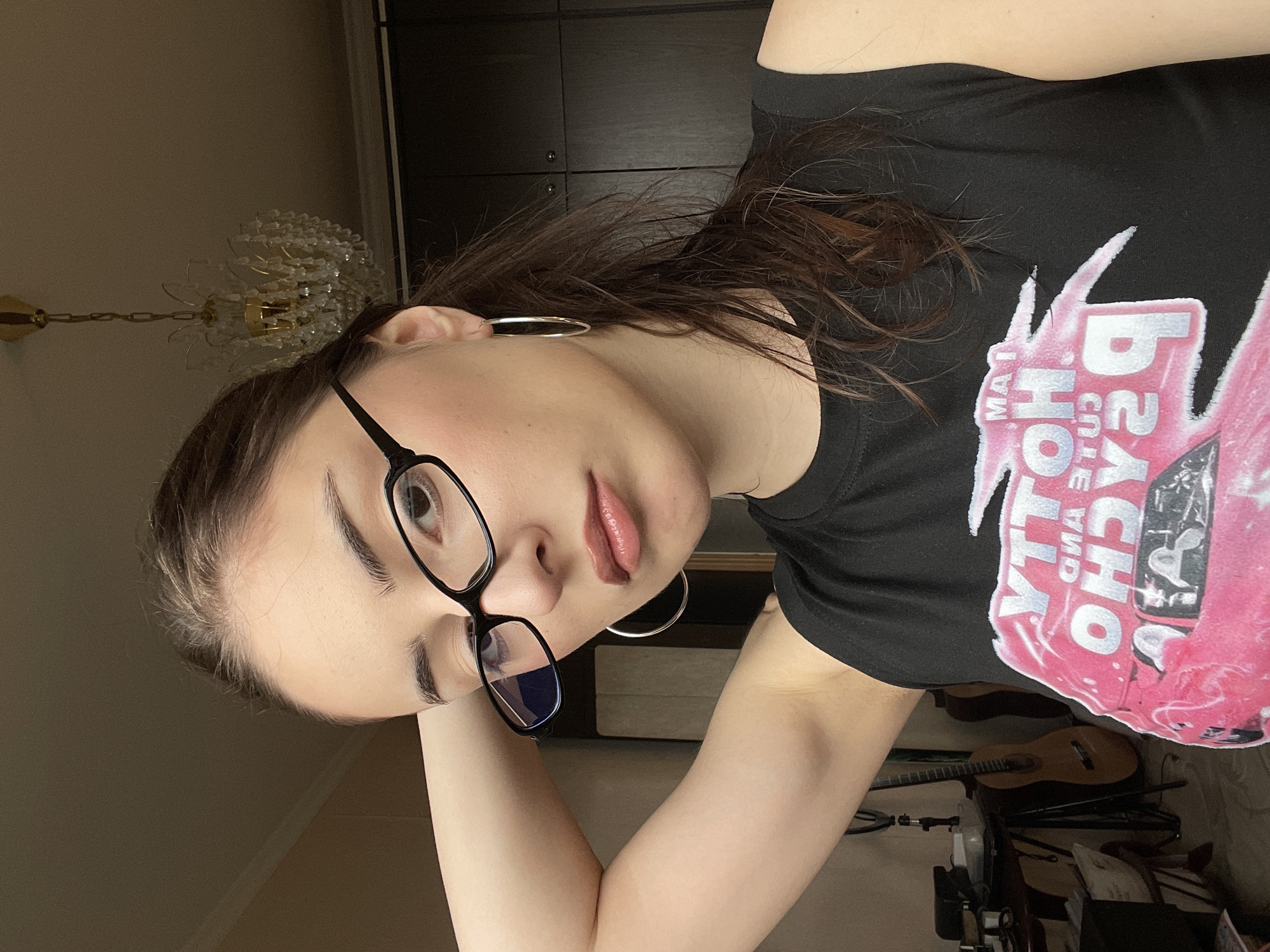 A young woman with brown hair, wearing black eyeglasses and hoop earrings, poses indoors. She is wearing a black t-shirt with a graphic print. Behind her are dark-colored cabinets, a chandelier, and a guitar. The setting appears to be a room, possibly in a home, with furniture and personal items visible in the background. There is no information to determine if the location is in Moscow, Russia.