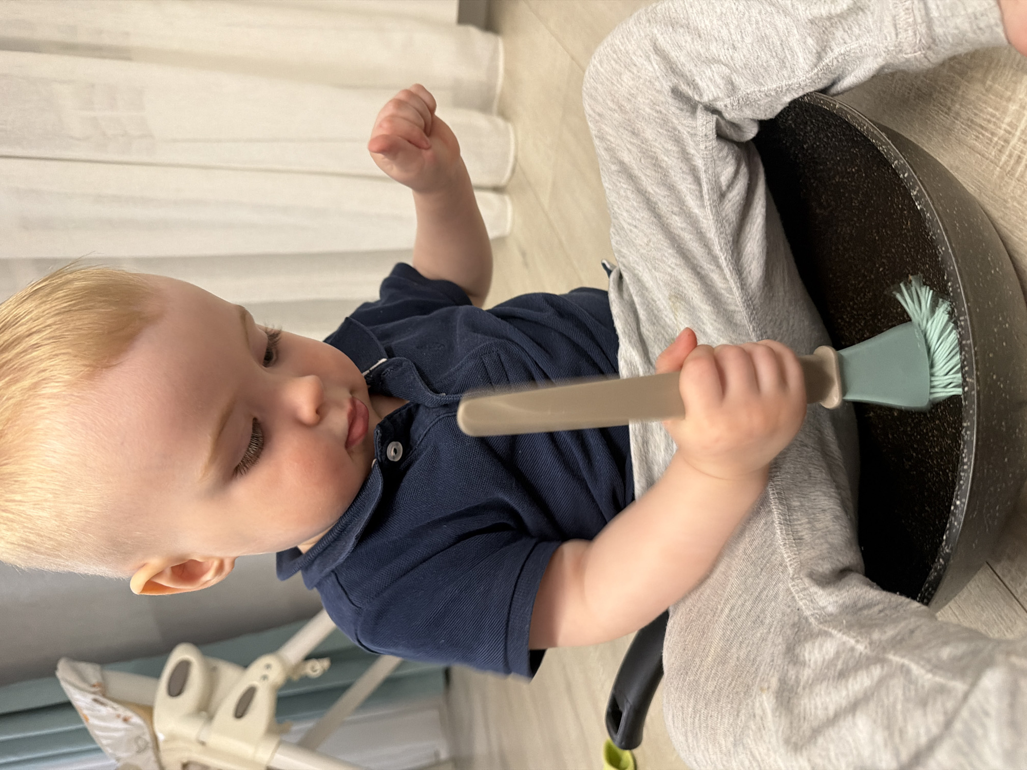 A child is sitting on a floor in what appears to be a domestic setting. The child, dressed in a navy blue polo shirt and light gray sweatpants, is holding a cooking utensil in their right hand and has a fist clenched in the air with their left hand. The child is seated in front of a dark-colored frying pan. The floor is light-colored, and a baby chair is visible in the background. The scene does not have sufficient data to place it in Votkinsk, Russia, specifically.