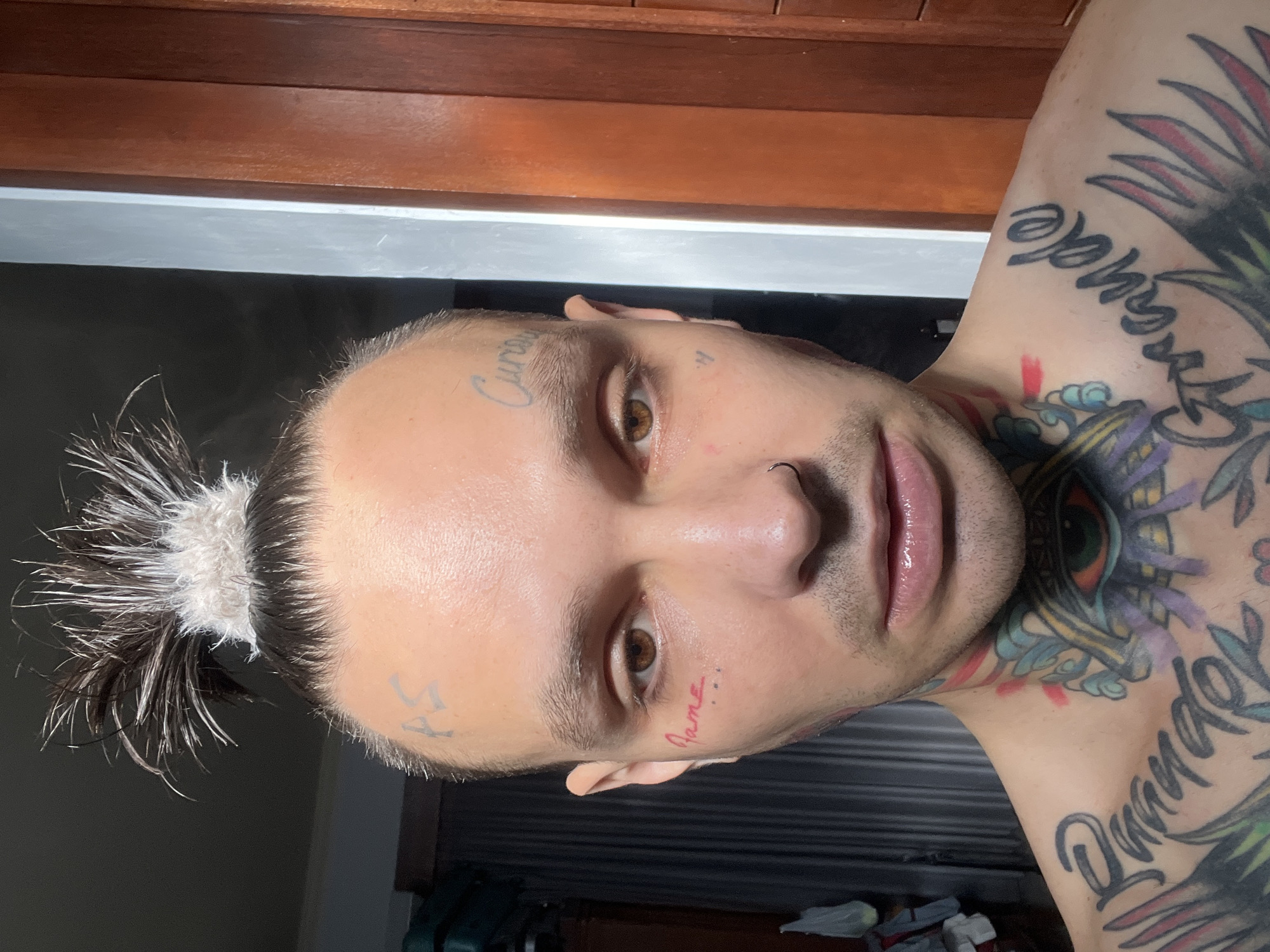 A close-up, frontal, medium-shot shows a fair-skinned person with dark hair pulled into a spiky topknot. The person has several tattoos on their face, neck, and chest. On the forehead, there are two tattoos: "PS" above the left eyebrow and "Curious" above the right. A small red tattoo that appears to say "Fame" is visible on the left cheek, near the eye. The person has brown eyes, a nose ring, and full lips. They are shirtless, revealing extensive colorful tattoos on their neck and chest, including an eye with rays emanating from it and script that reads "Grande" on the right shoulder. The background consists of a dimly lit interior with vertical blinds on the left and a wooden door on the right. The lighting is bright and comes from the front.