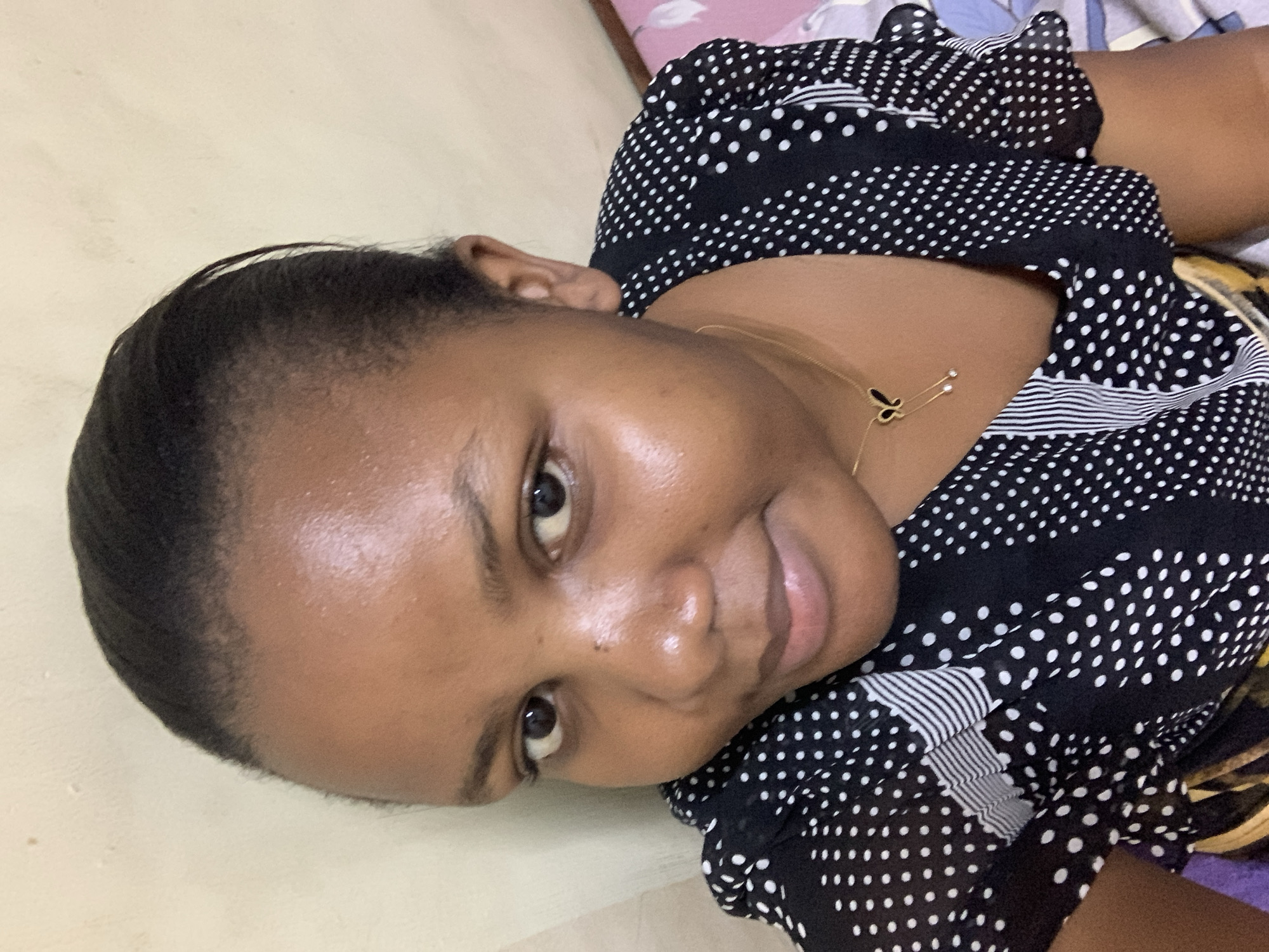 A medium, indoor shot of a woman with dark skin, possibly of African descent, facing the camera at an angle. She has short, dark hair neatly styled away from her face. She's wearing a black and white polka dot top with a small gold necklace. The background is a light beige wall on one side and bedding with a floral pattern on the other. There's no specific evidence in the image to confirm a location in Dar es-Salaam, Tanzania.