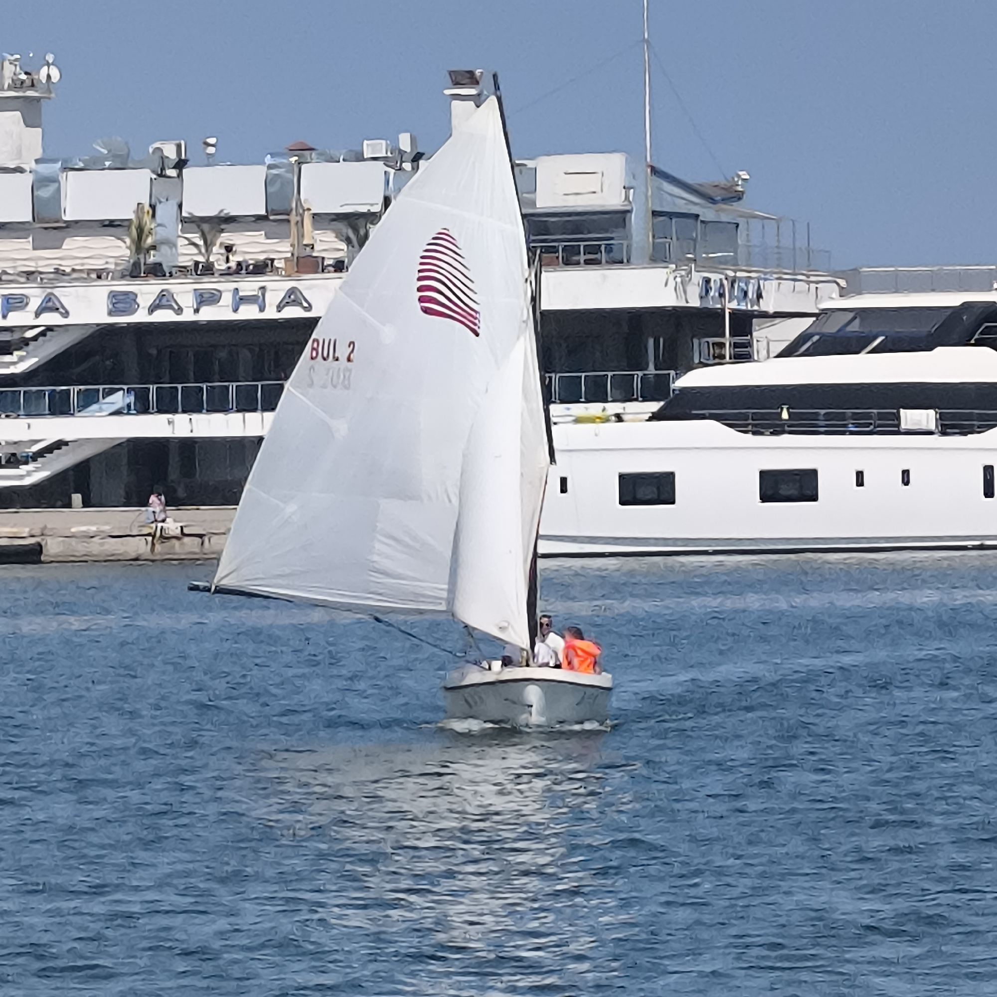 A white sailboat with a red and white striped insignia on its sail is navigating through the blue water of the Varna Bay in Bulgaria. The sailboat has two individuals on board; one is steering, and the other is seated at the rear wearing an orange life vest. In the background, a large white ship is docked, displaying the text "APA BAPHA" and "BUL 2" on its side. A person is visible on the dock in the distance, sitting on a wall. The sky above is clear and blue.