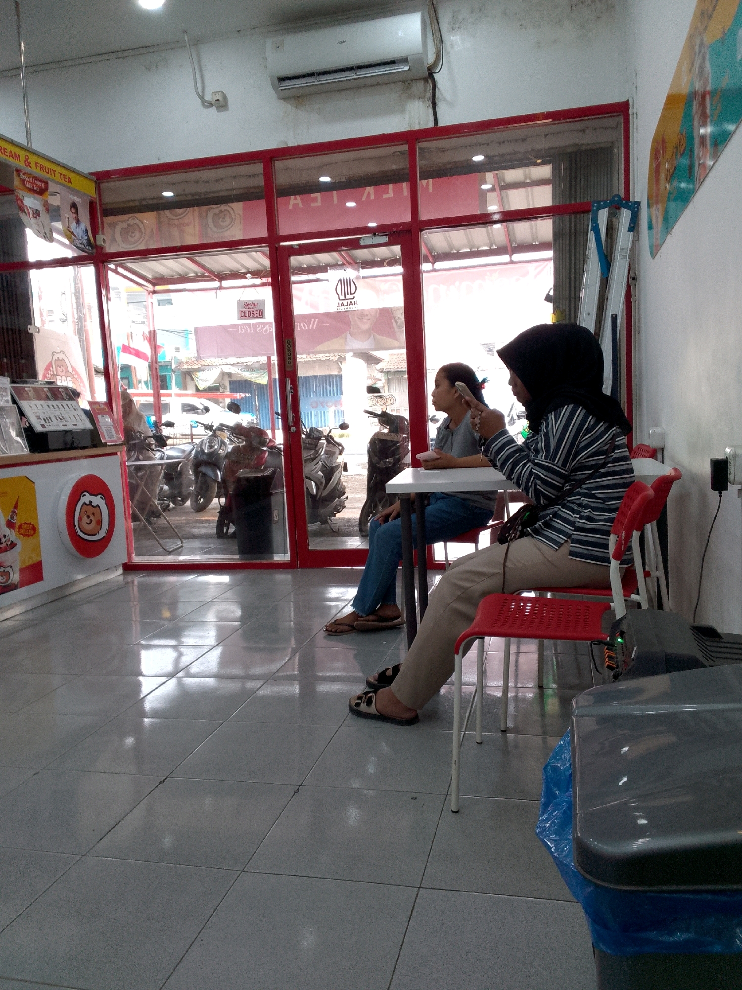 A medium shot captures the interior of what appears to be a food stall or shop in Rajamandala Kulon, Indonesia. The foreground is dominated by a gray tiled floor, reflecting the overhead lighting and the interior of the establishment. To the left, a counter displays various items, including what looks like an ice cream cone and informational pamphlets. A red and white circular logo with a cartoon bear face is prominently featured on the counter.
In the mid-ground, two women are seated at a white table. The woman on the right wears a black hijab and a striped long-sleeved shirt, with khaki pants. She is holding a smartphone in her right hand and looking down at it. The woman on the left, wearing a gray t-shirt and blue jeans, is looking towards the right and appears to be holding a phone or similar object. She is seated on a red chair.
Behind the women, through a large glass window framed in red, is an outdoor scene. Several motorcycles are parked in what seems to be a street or parking area. Above the motorcycles, the entrance to buildings with awnings and signs is visible. A sign with the word "CLOSED" in red letters is hanging on the glass door.
The environment suggests a commercial area, possibly a roadside eatery or a small business catering to locals. The presence of motorcycles indicates a mode of transportation common in the region. The overall scene is casual and everyday, depicting a moment of pause or waiting within a business setting. The lighting is bright and somewhat harsh, highlighting the reflective surfaces of the floor and the interior of the shop.