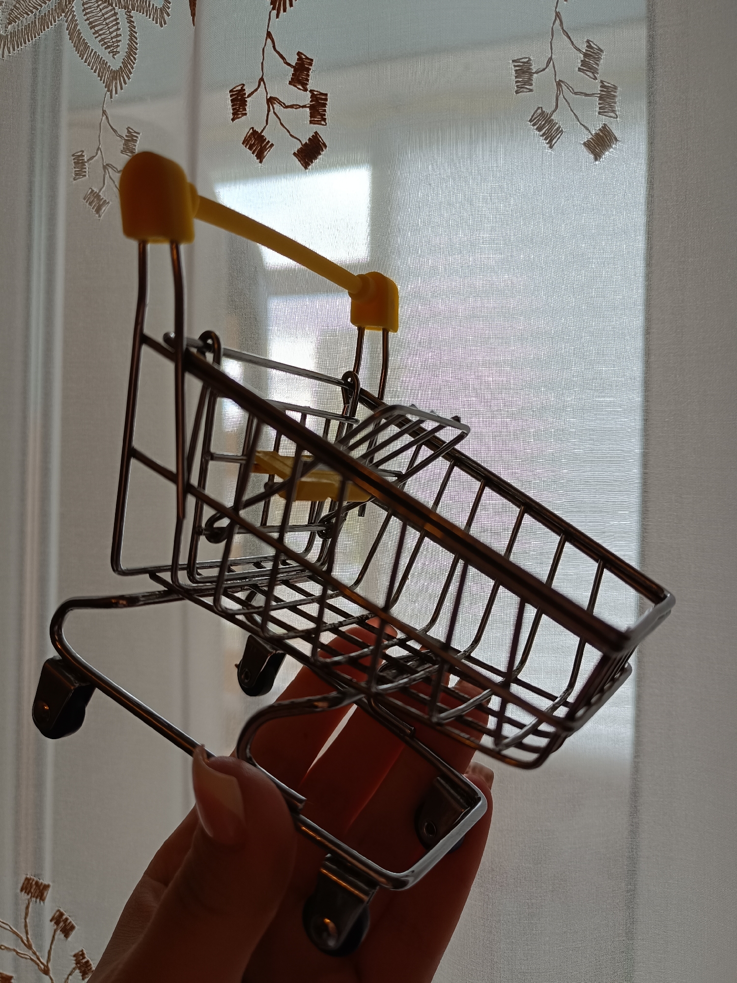 A person holds a miniature shopping cart in their hand against a backdrop of a white, patterned curtain. The shopping cart is made of a metallic wire frame with a yellow handle. The curtain has a floral embroidery pattern. The location is not identifiable as Tobolsk, Russia, based on the provided image.
