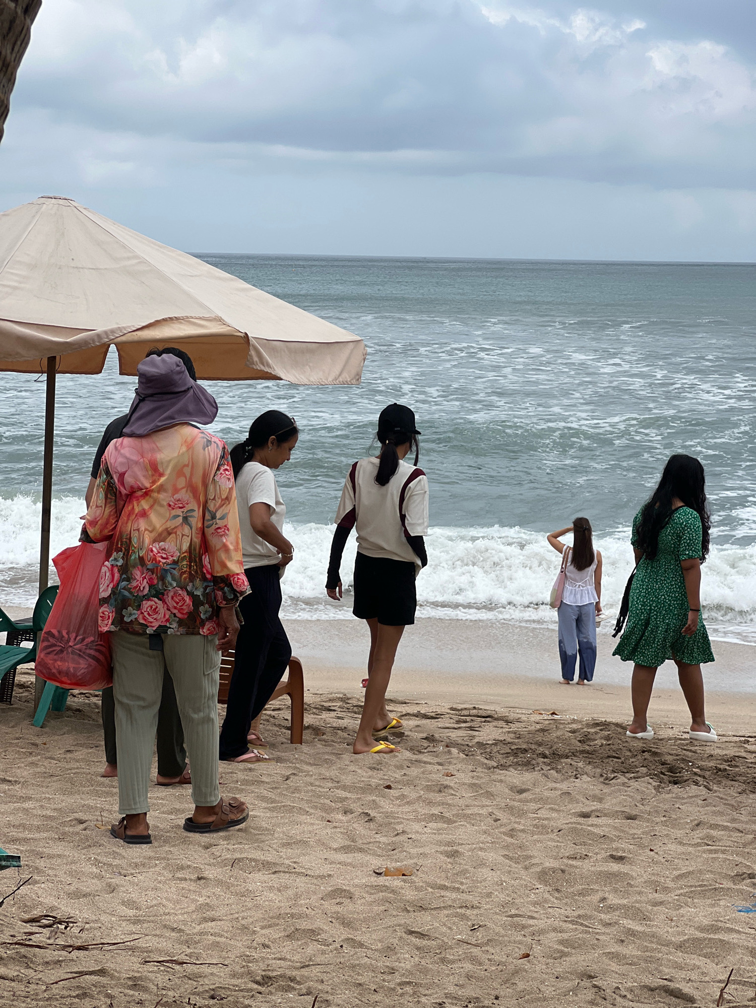The image depicts a beach scene in Kuta, Indonesia, under an overcast sky. Several people are present on the sandy shore, with the ocean waves breaking in the background.
In the foreground, a person wearing a wide-brimmed hat and a brightly colored floral shirt with green pants stands near a large, light-colored umbrella. They are holding a red plastic bag. To their right, another individual in a white t-shirt and dark pants is partially visible, facing away from the camera. Further to the right, a person in a black cap and a white and maroon athletic top with black shorts stands facing the ocean.
In the middle ground, a woman in a white top and blue jeans stands facing the ocean, with her hand on her head. To her right, a person in a green patterned dress stands facing away from the camera, looking towards the water.
The environment consists of a sandy beach with some scattered debris and vegetation. The ocean is visible with moderate waves. There are beach chairs and parts of umbrellas scattered on the sand. The overall impression is that of a somewhat cloudy day at a popular beach location.