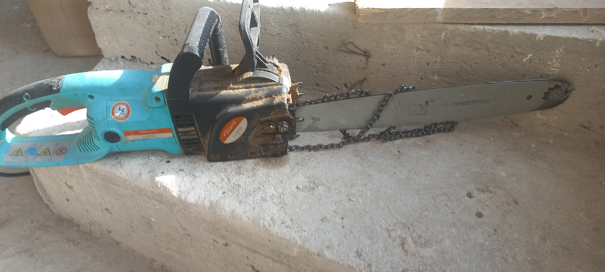 The image displays an electric chainsaw resting on a concrete surface. The chainsaw is predominantly turquoise and black, with a metal bar and chain. The brand "Sturm" is visible on the side of the chainsaw. The surrounding environment appears to be an outdoor or construction area, characterized by rough concrete textures and some debris. The lighting suggests it is daytime, with shadows cast across the scene. There are no people present in the image, and no discernible actions or interactions are taking place. The scene is static and presents the chainsaw as the primary subject. The specific location of Dagestanskiye Ogni, Russia, is not visually evident from the image content.