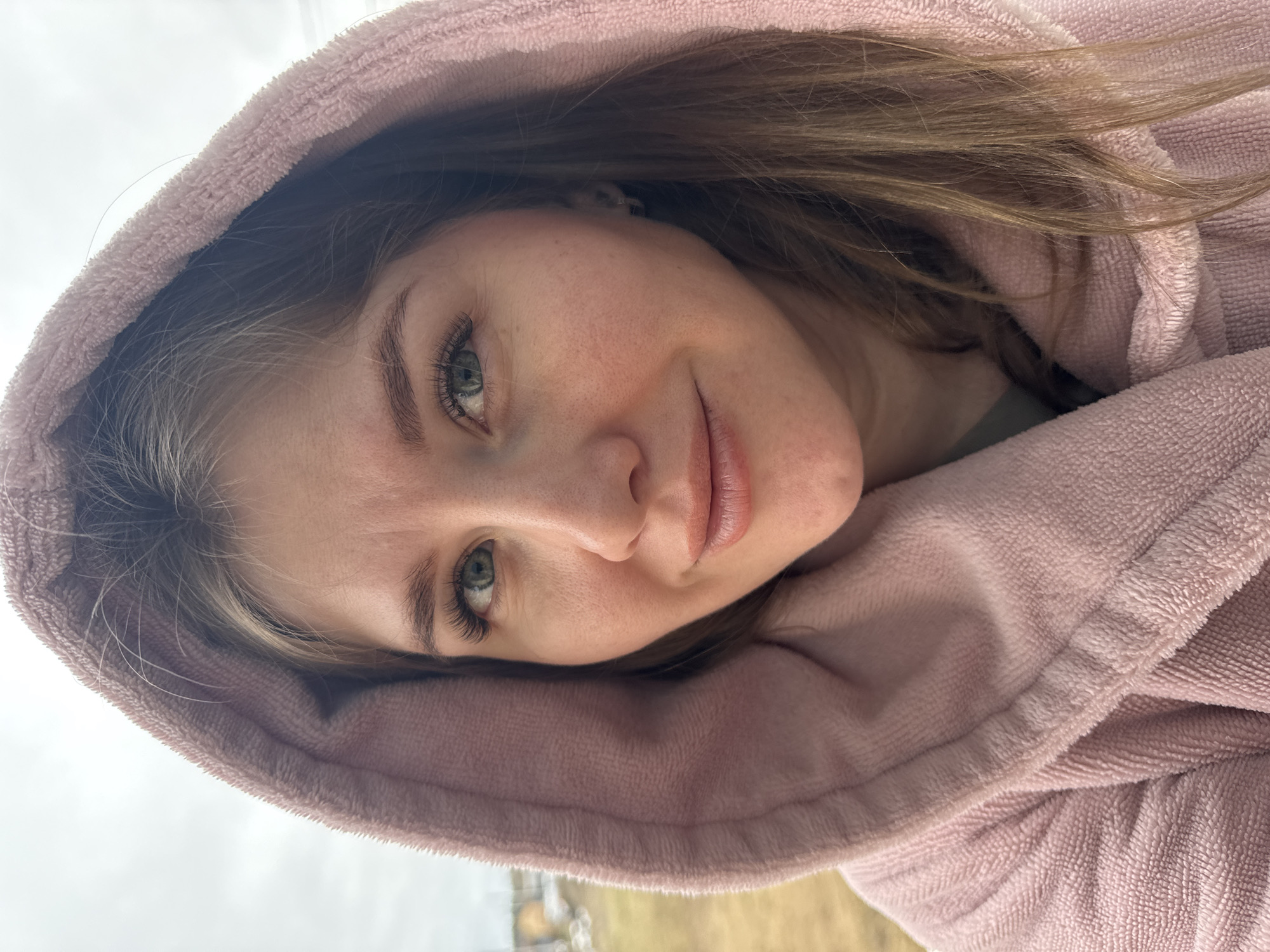 A close-up, slightly angled portrait of a young woman with fair skin and light brown hair, wearing a pink hooded robe. Her gaze is directed towards the camera with a subtle smile. She has green eyes and visible eyelashes. The background appears to be an outdoor setting with muted tones, possibly depicting a landscape. The exact location is Gornyi (Dvurechenskoe S/P), Russia, as specified.