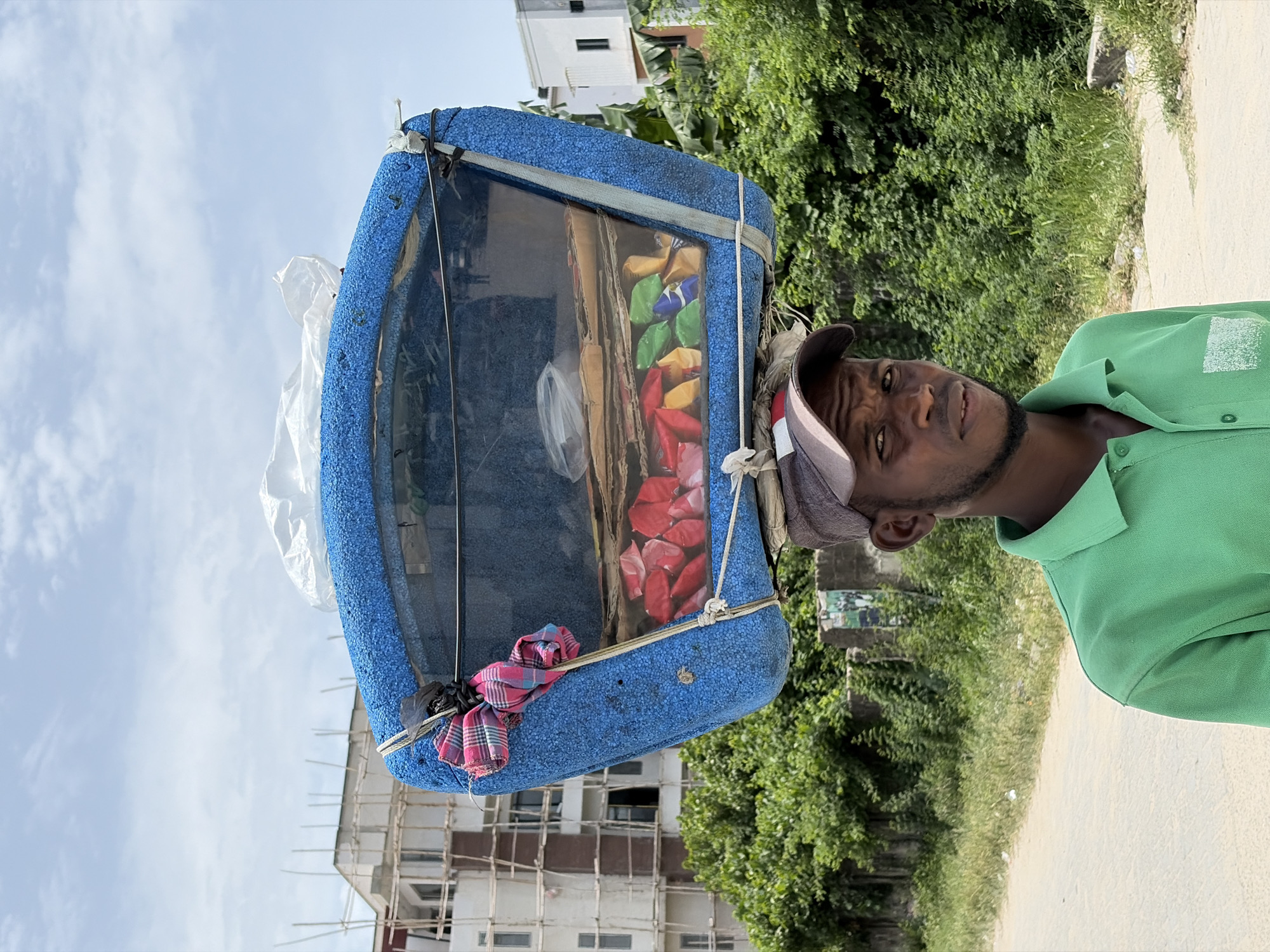 In Igboefon, Nigeria, a man stands on a roadside wearing a green shirt and a cap. He is carrying a blue, framed container on his head, which displays colorful wrapped items. Behind him are buildings, trees, and a clear sky.