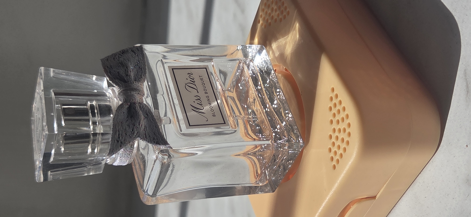 The image shows a close-up, slightly elevated view of a rectangular glass perfume bottle, identified by a label as "Miss Dior Blooming Bouquet." The bottle is clear and filled with a pale pink liquid. A textured grey bow is tied around the neck of the bottle, just below a faceted clear cap. The bottle rests on a peach-colored object with a textured surface featuring a pattern of small, circular indentations. The background consists of a light-colored surface with dappled shadows, suggesting sunlight is illuminating the scene. The overall impression is one of a still life arrangement, possibly a product photograph or a personal item displayed. There are no people or indications of the city of Karaganda, Kazakhstan, in this image.
