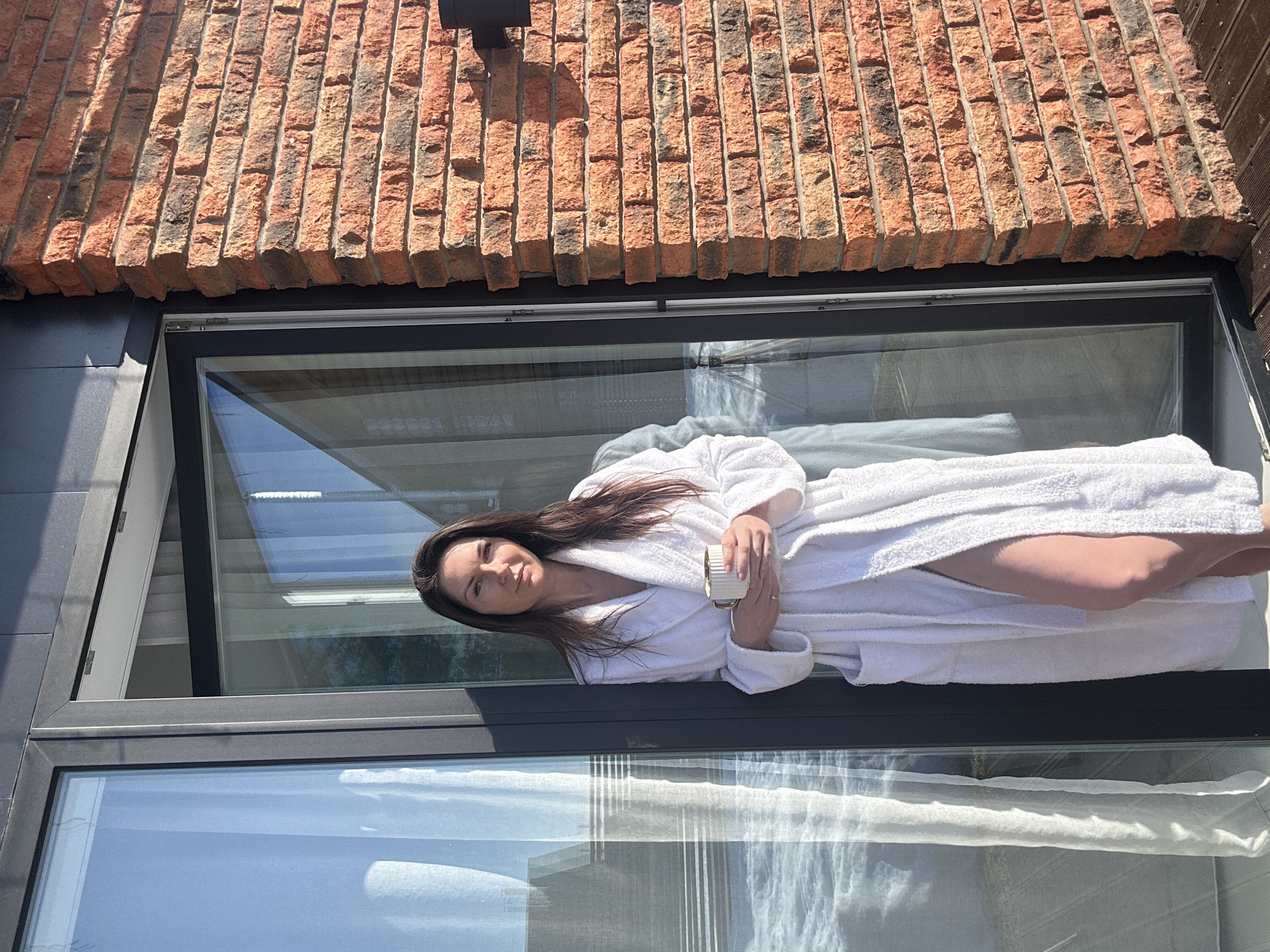 A female subject is depicted standing in a partially open dark-framed glass doorway, facing forward in Chaikovsky, Russia. She has dark, shoulder-length hair and wears a white terry-cloth bathrobe, which is open to reveal her right thigh. She holds a white and gold mug with both hands at chest level. To her right, a wall of reddish-brown brick with dark mortar is visible, featuring an outdoor wall light fixture. A dark, slatted wooden deck forms the ground surface at the bottom right. Through the glass panel to her left, reflections show a modern building with an angled roofline and what appears to be a snow-covered or frozen expanse of water. The scene is under bright natural sunlight, casting distinct shadows.