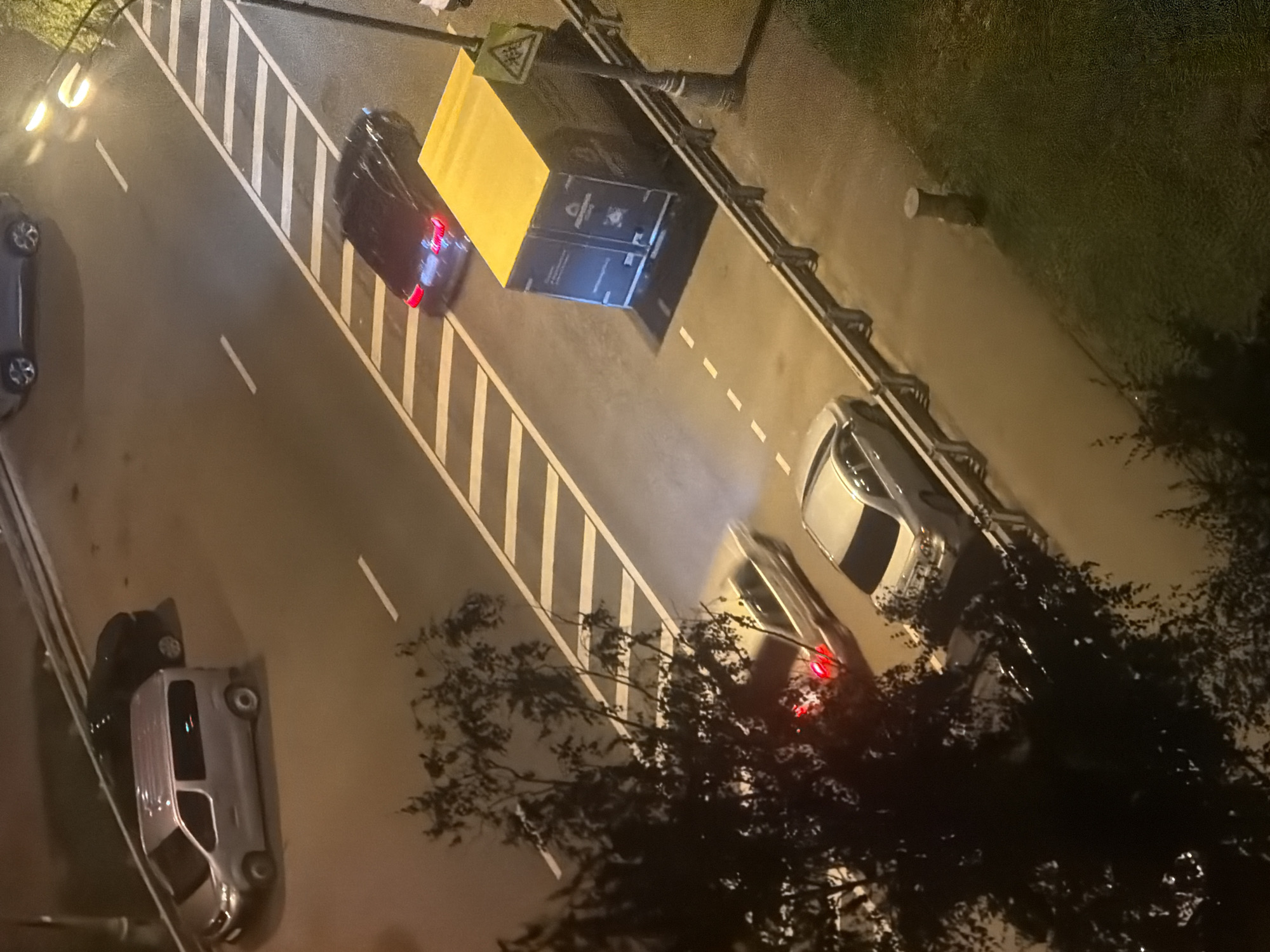 The image shows a street scene at night in Moscow, Russia. Several vehicles are visible, including a silver minivan, a black sedan, a silver sedan, and a truck with a yellow cargo box. The vehicles are on a wet road with white lane markings. There is a pedestrian crossing with white stripes. A street lamp is visible, and trees line the side of the road. The scene appears to be in a residential or mixed-use area.