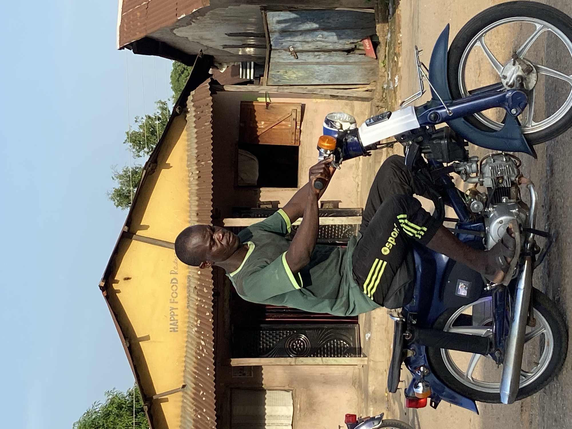 A man is sitting on a blue motorcycle in front of a yellow building. The building is labeled "HAPPY FOOD Res" (presumably a restaurant). The man is Black, wearing a green t-shirt with yellow trim, black shorts with yellow stripes, and sandals. He is holding the handlebars of the motorcycle. The building behind him has a corrugated metal roof and a dark door. The environment appears to be a street in Mararaba, Nigeria.