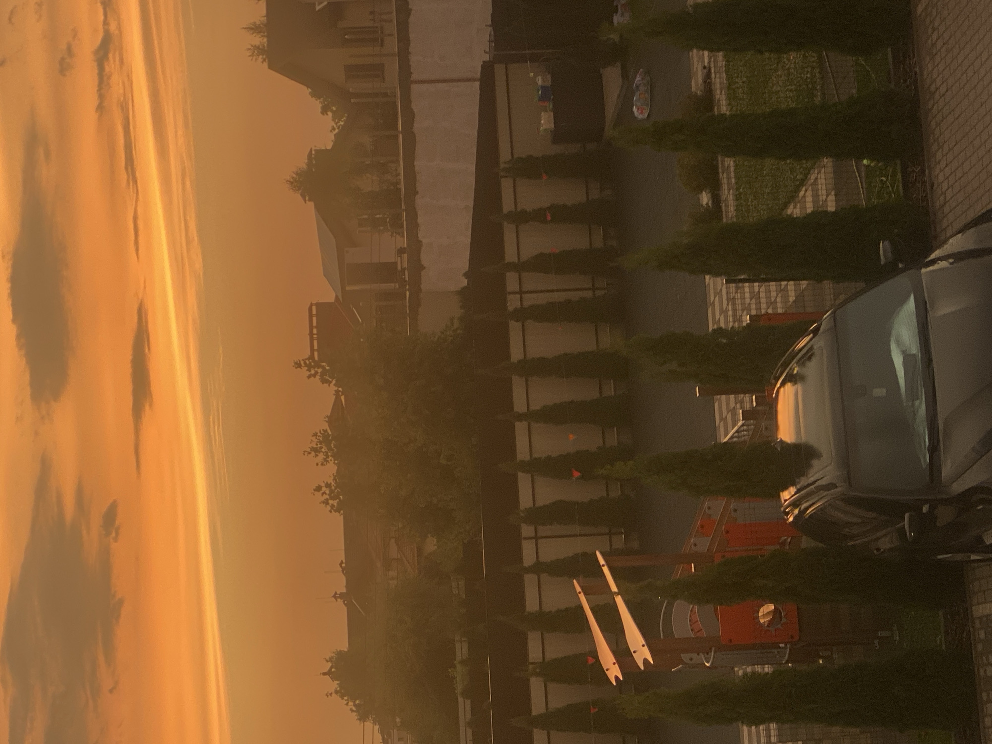 The image depicts a suburban scene in Novi Petrivtsi, Ukraine, at dusk. The sky is filled with vibrant orange and yellow hues, indicating a sunset with scattered clouds. In the foreground, a dark-colored SUV is parked on a paved driveway. Adjacent to the car, a row of tall, conical evergreen trees lines a fence. Beyond the trees and fence, a playground structure with orange and white elements is visible. In the background, houses with visible rooftops and trees are silhouetted against the colorful sky, suggesting a residential neighborhood. The overall atmosphere is peaceful and serene, characteristic of a quiet evening in a semi-rural setting.