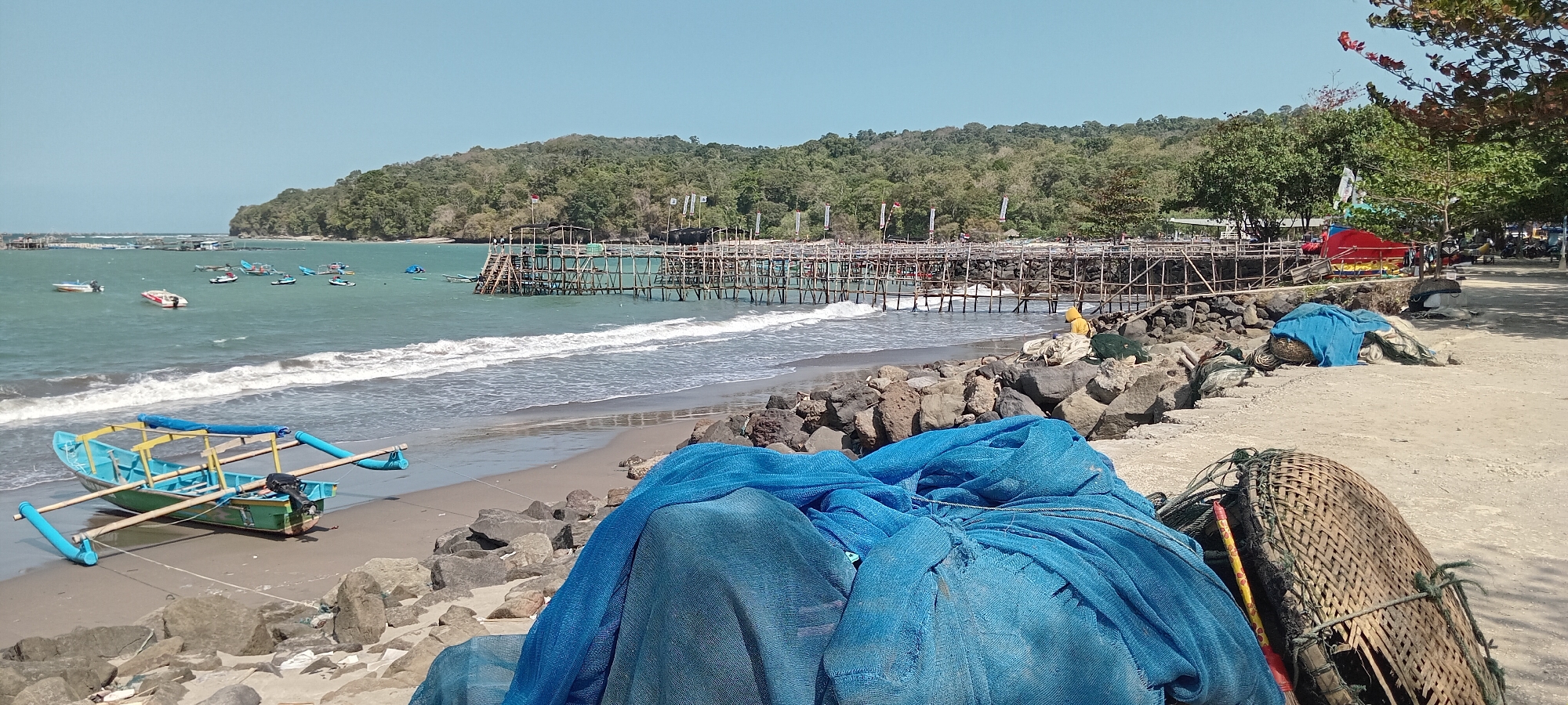 The image depicts a beach scene in Desa Pagandaran, Indonesia, on a clear, sunny day. The foreground is occupied by a pile of blue fishing nets and a woven basket. To the left, a small, light blue and green boat with outriggers rests on the sandy shore, a motor attached to its stern. The beach is rocky, with large stones scattered along the waterline. In the mid-ground, the turquoise ocean has moderate waves breaking on the shore. Several small boats are visible further out in the water. Dominating the mid-ground is a long wooden pier or jetty made of bamboo, extending into the sea. The pier has a walkway with stairs leading down to the water and is supported by numerous wooden stilts. Some figures can be seen on the pier. In the background, a lush green, tree-covered hill rises above the pier and the coastline. The sky is clear and blue. To the right of the pier, on the sandy area adjacent to the beach, there are more fishing nets, some red and yellow structures, and a few motorcycles parked under the shade of trees.