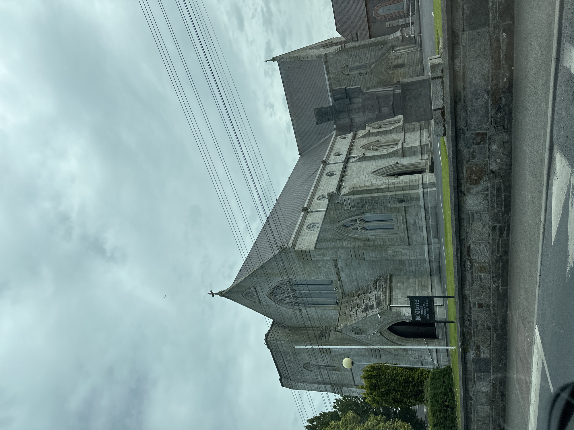The image shows the exterior of St. Clare's Church in Carlow, Ireland. The church is constructed of grey stone with Gothic architectural elements, including pointed arch windows and a prominent cross on the roof. There is a sign in front of the church that reads "ST CLARE'S MASS TIMES" followed by a schedule of services. Power lines are visible overhead. The foreground shows a stone wall and a road with a crosswalk. The sky is overcast and cloudy.