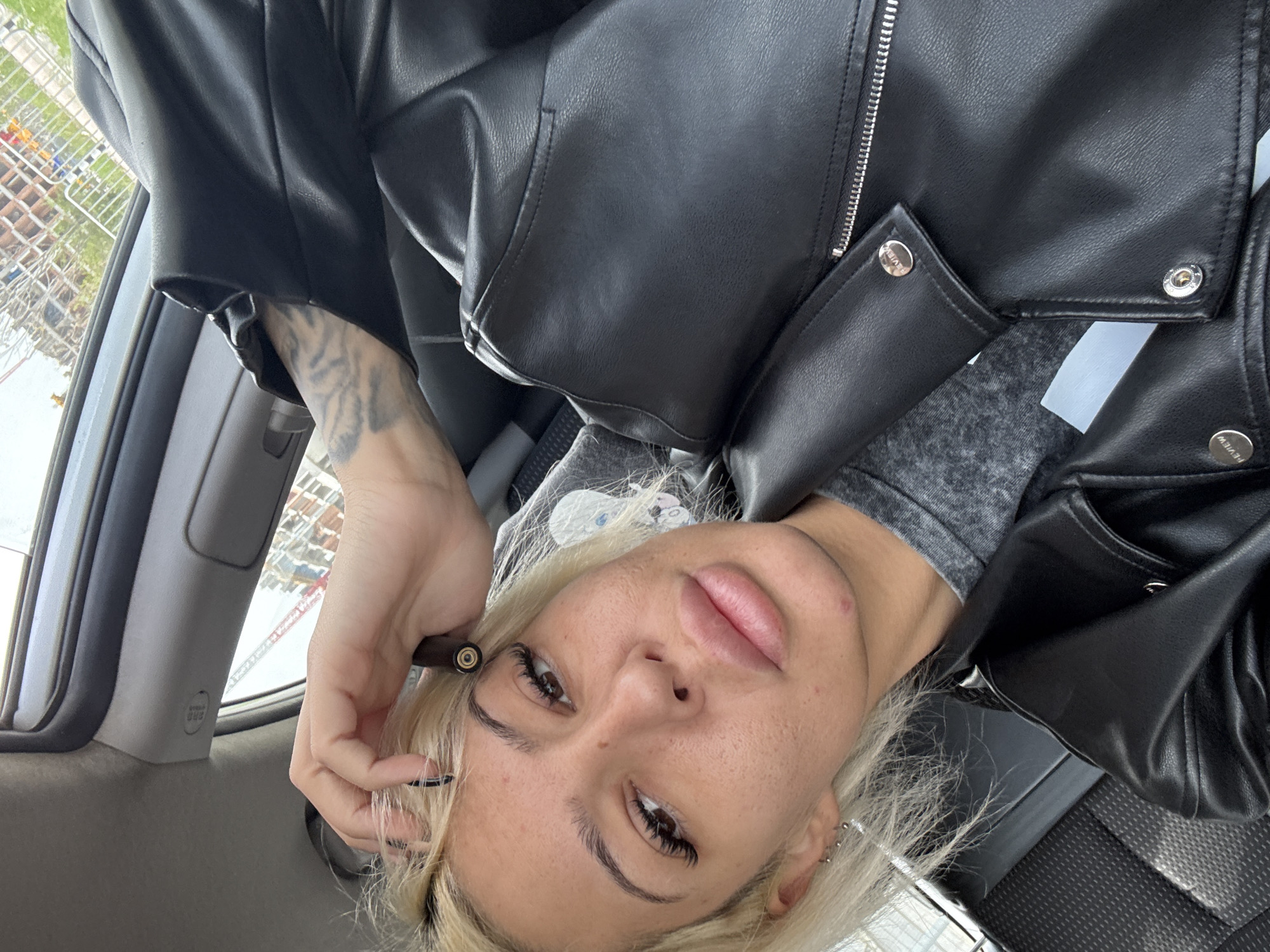 A person with blonde hair is sitting in a car wearing a black leather jacket and a gray long-sleeved shirt. The person is holding a vape pen with their right hand near their face. The background shows a partial view of the car's interior and a cityscape seen through the window. The image appears to be taken at an unusual angle.