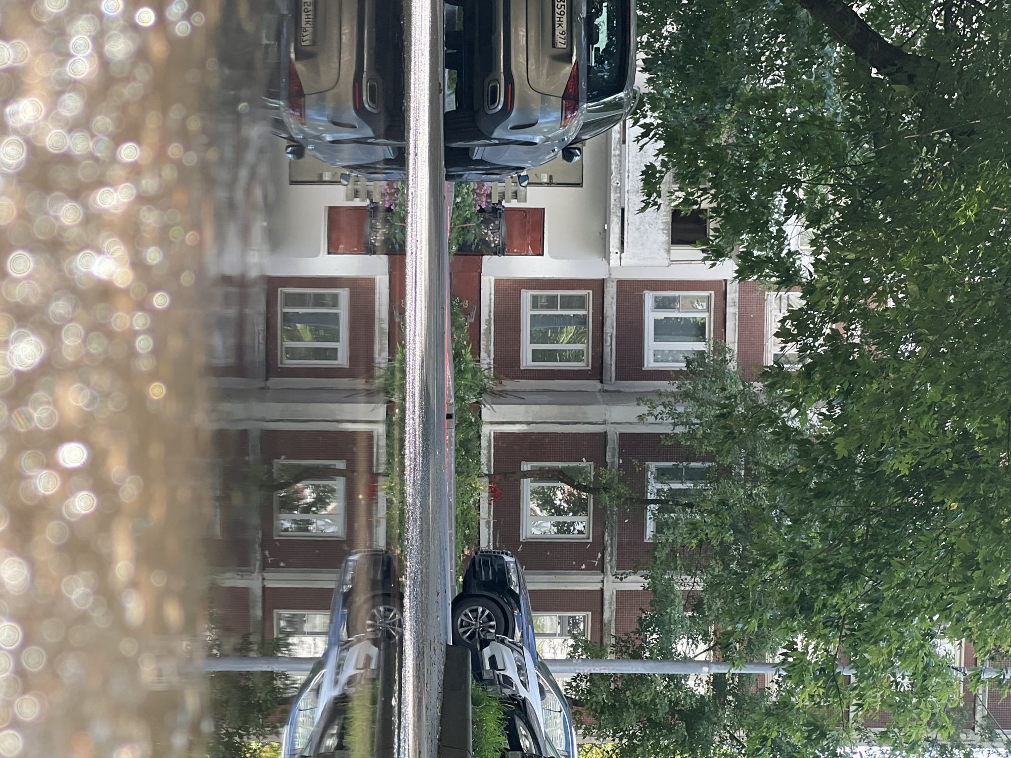 The image depicts a residential building in Moscow, Russia, with a focus on the reflections in a puddle on the ground. The building appears to be a typical Soviet-era apartment block, with a red brick facade and white window frames. The building is surrounded by trees, and there are several cars parked in front of it. The puddle in the foreground reflects the building, the trees, and the cars, creating a distorted and dreamlike image. The overall impression is one of a quiet, ordinary day in a Russian city.