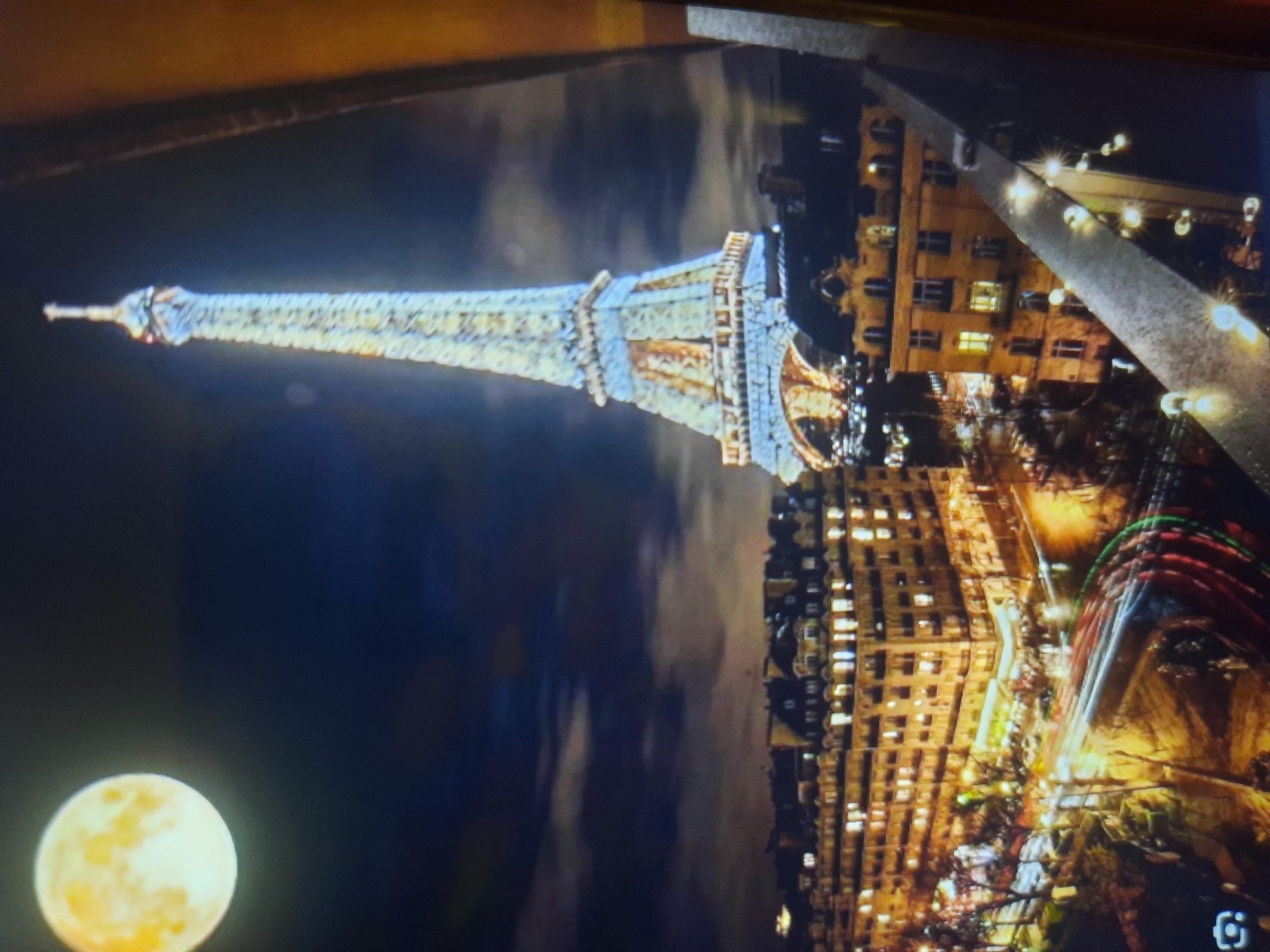 The image displays a nighttime scene of the Eiffel Tower, illuminated and set against a dark, cloudy sky with a large, bright moon in the upper left quadrant. Below the tower are Parisian-style buildings with glowing windows, indicating urban activity. Streaks of light from moving vehicles are visible on the streets, suggesting traffic. The foreground shows a metal railing adorned with small, decorative lights. The overall composition is a wide shot capturing a vibrant cityscape at night. This scene is of Paris, France, not Rosenheim, Germany.