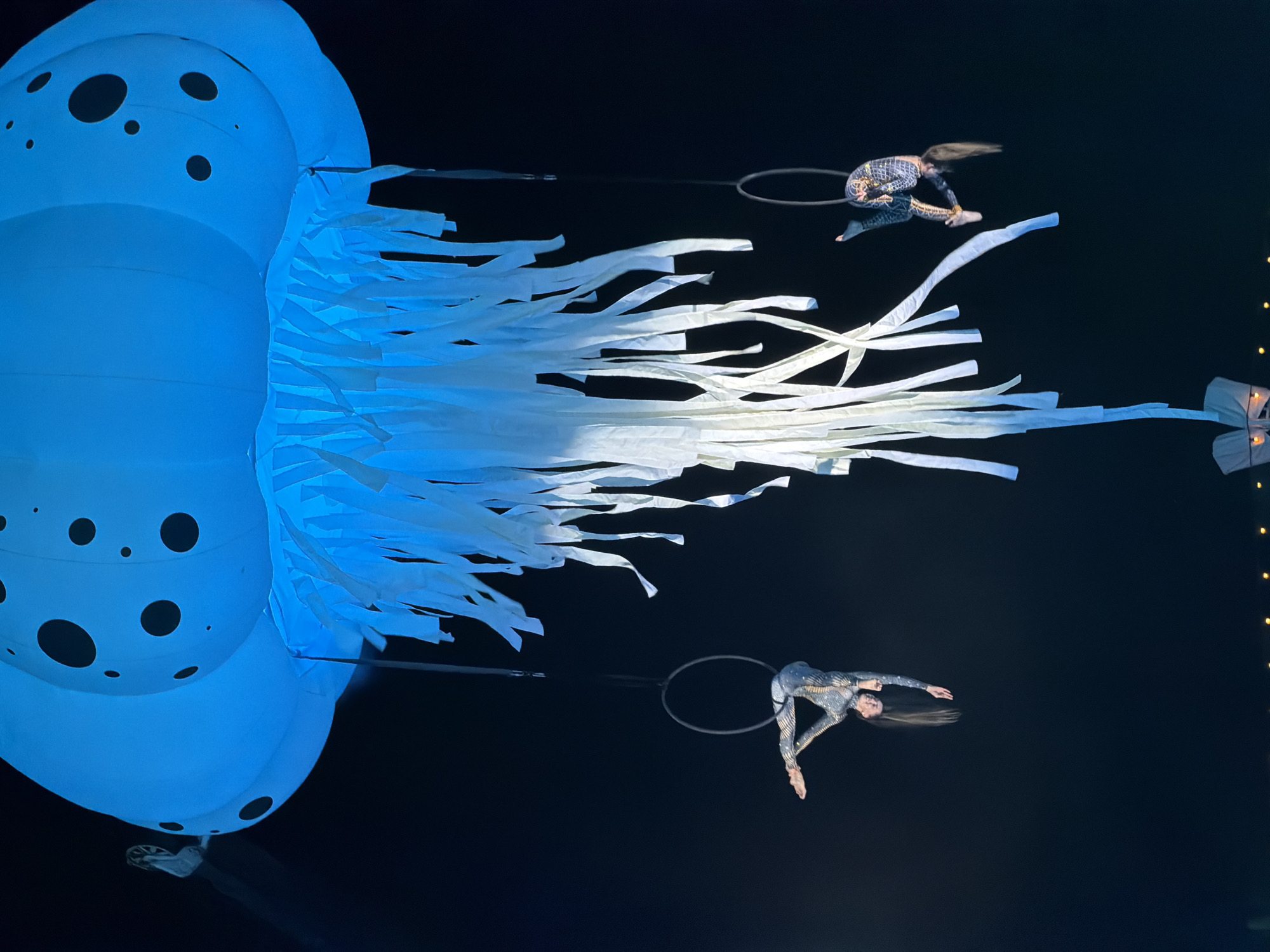 An aerial performance is taking place under a large, glowing blue jellyfish-shaped inflatable. The jellyfish has black polka dots and long, white fabric streamers hanging from its bottom. Two performers, dressed in sparkling, form-fitting costumes, are suspended in the air on aerial hoops. The performer on the left is positioned upside down, with her legs bent and arms extended. The performer on the right is also inverted, with one leg tucked and the other extended, and her arms are bent. The background is dark, suggesting it is nighttime. The scene appears to be an outdoor performance in Odesa, Ukraine, with decorative lights visible in the lower part of the frame, hinting at a public event or festival.