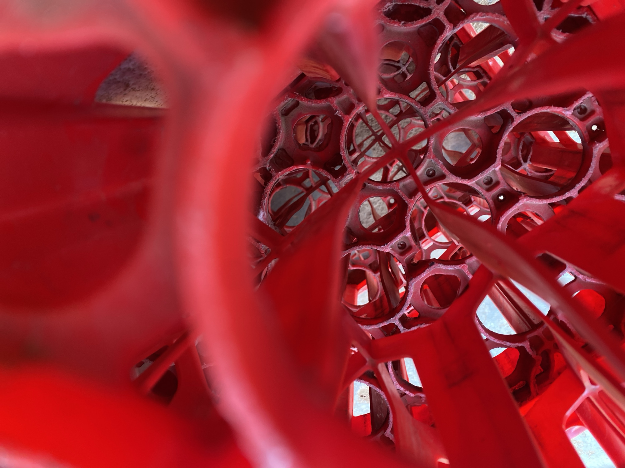 The image is a close-up, abstract view looking into a red plastic container with numerous circular openings. The container's structure creates a repeating pattern of interconnected circles and struts. The exterior of the container is out of focus, while the interior structure is partially in focus. The material appears worn, with some surface imperfections visible. Background elements visible through the holes suggest an exterior environment. There are no people visible. The image does not provide enough information to determine the location as Maigizo, Nigeria.
