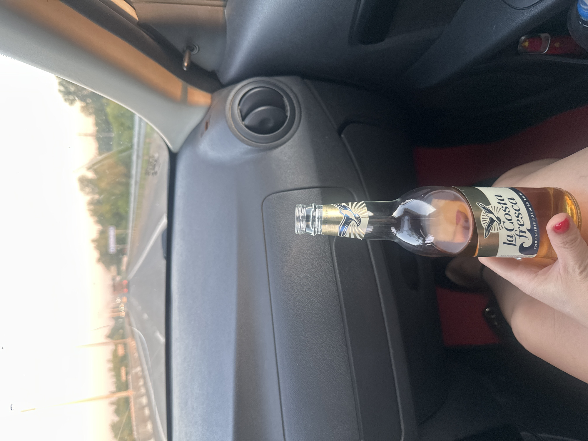 A hand holds a bottle of "La Costa Fresca" beer while riding in a car on a highway at sunset. The car's dashboard and a portion of the passenger seat are visible. The highway appears to be near Novoaltaysk, Russia, with trees and streetlights lining the road. The sky is hazy with the warm colors of sunset. The bottle is partially filled with a light-colored liquid and has a golden label with a bird graphic.