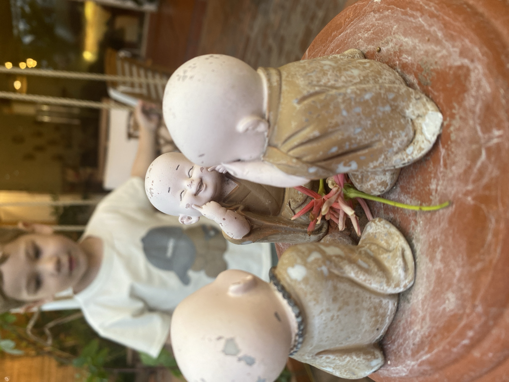 The image, possibly taken in Phu Thuy Ward, Vietnam, shows three small monk figurines arranged in a circular pattern on what appears to be a terracotta-colored planter. The figurines are made of a light-colored material with details like robes and heads. In the background, a child wearing a white t-shirt with a gray cloud emblem is partially visible. Behind the child, there's a blurred view of a building with a reflection in the window. Some greenery is also visible at the bottom of the frame. The figurines are grouped closely together.