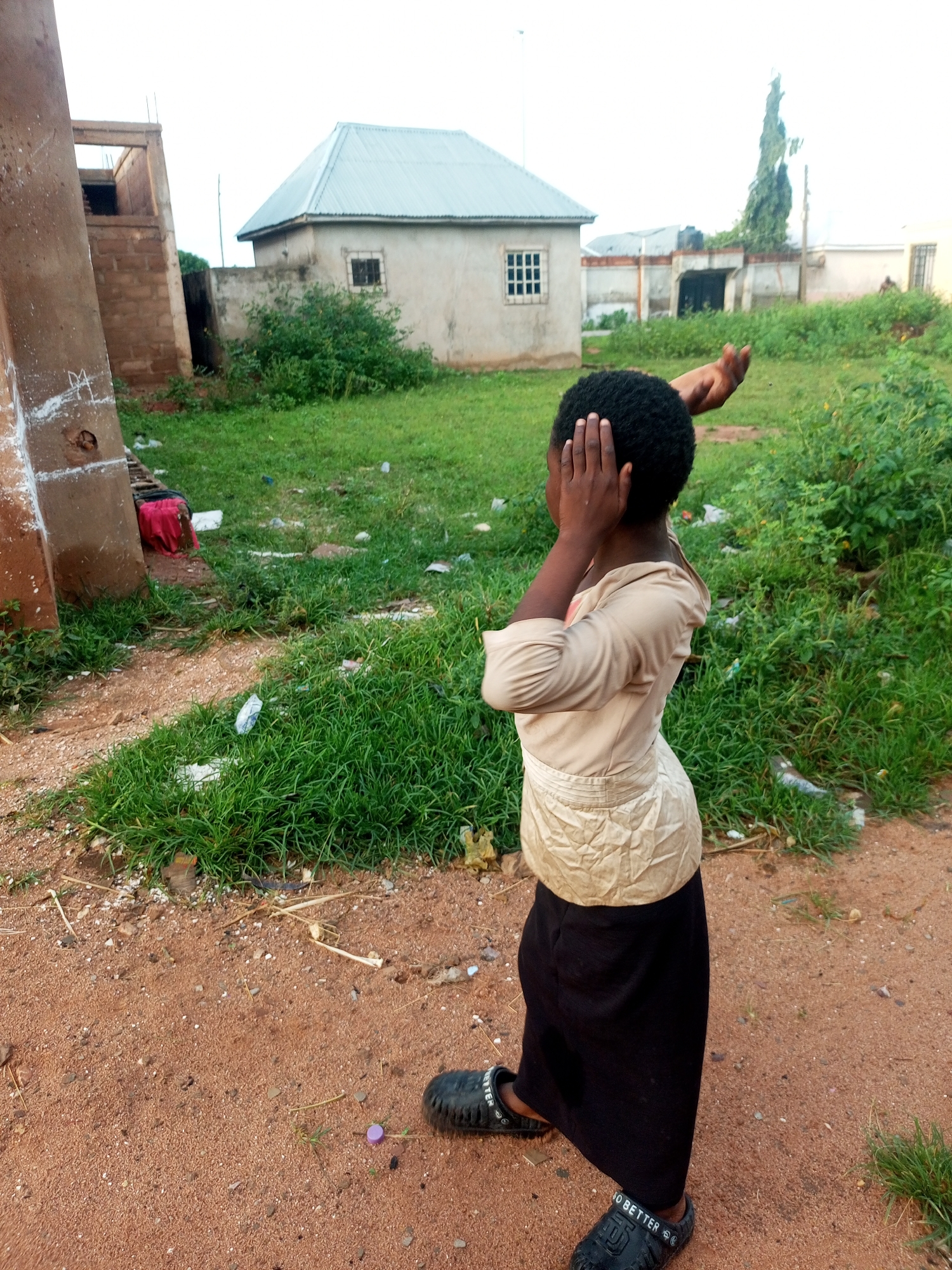 In Tempure, Nigeria, an outdoor scene depicts a young person standing on sandy ground. The individual is facing away from the camera, wearing a light-colored, long-sleeved shirt and a long black skirt. Their hands are covering their ears, and their right arm is raised. The environment appears to be a somewhat unkempt area with patches of green grass and scattered debris. In the background, there are several buildings, including a small house with a silver corrugated roof and a partially constructed building made of brick. The sky is overcast.