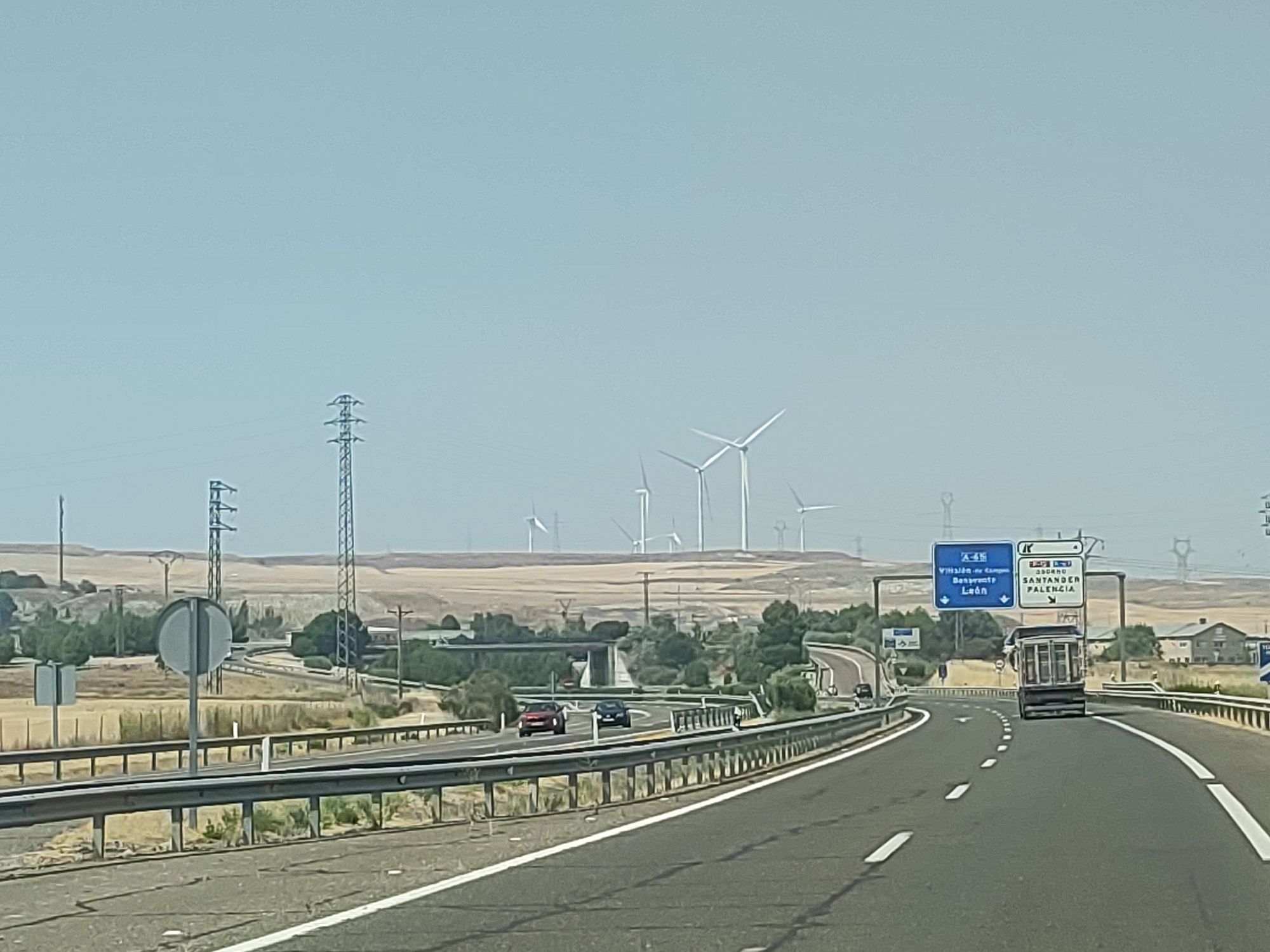 The image depicts a view from a highway in Palencia, Spain. A truck is visible in the foreground, driving on the right side of the road. In the distance, a field of wind turbines can be seen, along with power lines and pylons. The sky is clear and blue. A highway sign indicates directions to Santander and Palencia, confirming the location. Cars are also driving on the highway. The landscape appears to be arid and rural.