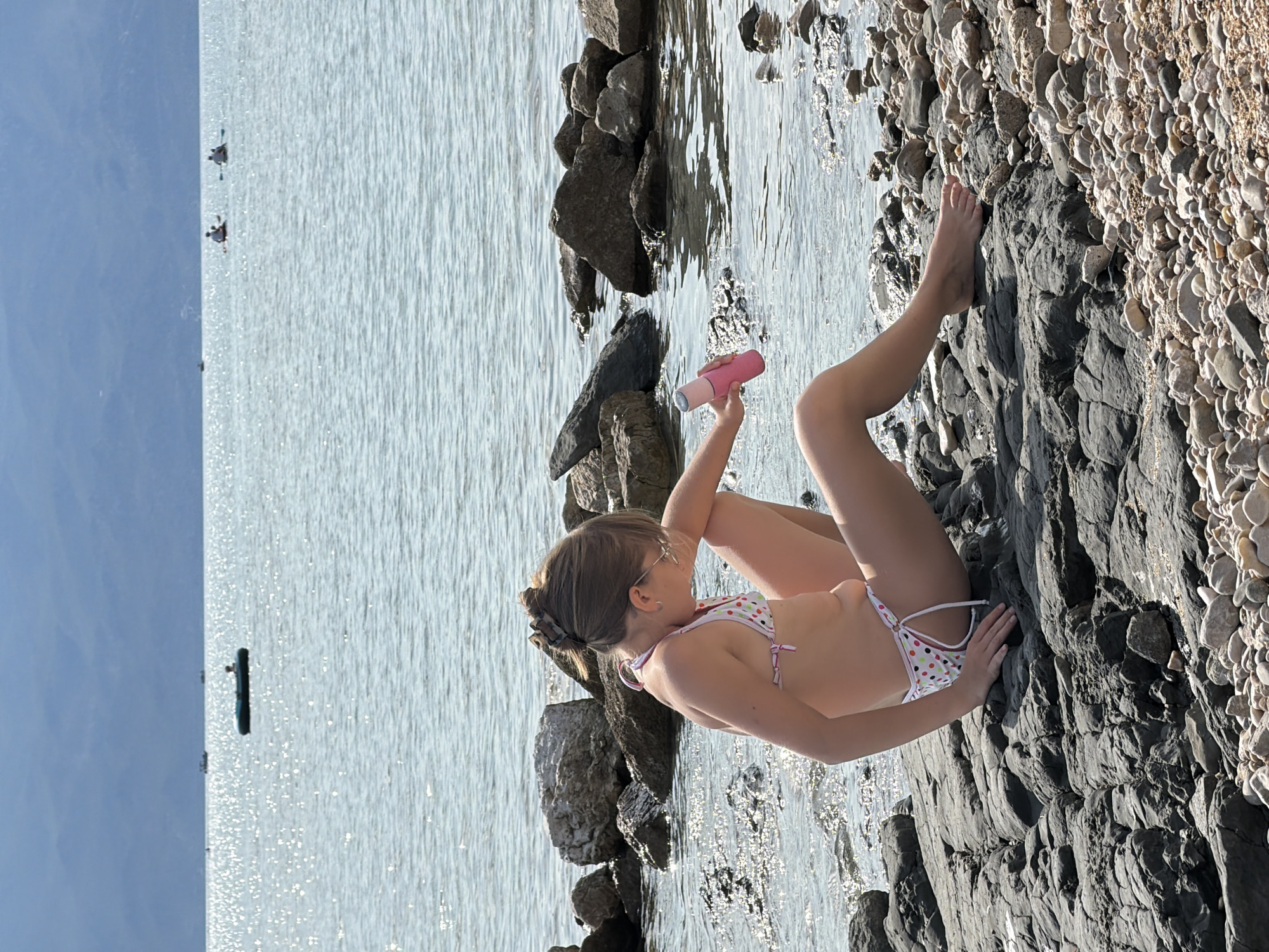 This image depicts a young girl in a polka-dot bikini sitting on a rocky shore near the water. She is holding a pink bottle in her right hand, and her left hand rests on the rocks beside her. Her legs are extended, with her right leg bent at the knee. The water appears calm with sunlight reflecting off its surface, suggesting a clear day. Behind her, there are more large rocks and a vast body of water that stretches to the horizon, where a faint blue sky is visible. In the distance, there are a few small boats or kayaks on the water, and a dark object that could be a buoy or a small vessel. The ground she is sitting on is a mix of dark, layered rocks and smaller, lighter pebbles. The scene appears to be set during daylight hours, likely in the afternoon given the sunlight. The girl's posture and gaze suggest a moment of relaxation or contemplation. The location context provided is Općina Baška, Croatia, which aligns with the coastal scenery. There is no visible text in the image.