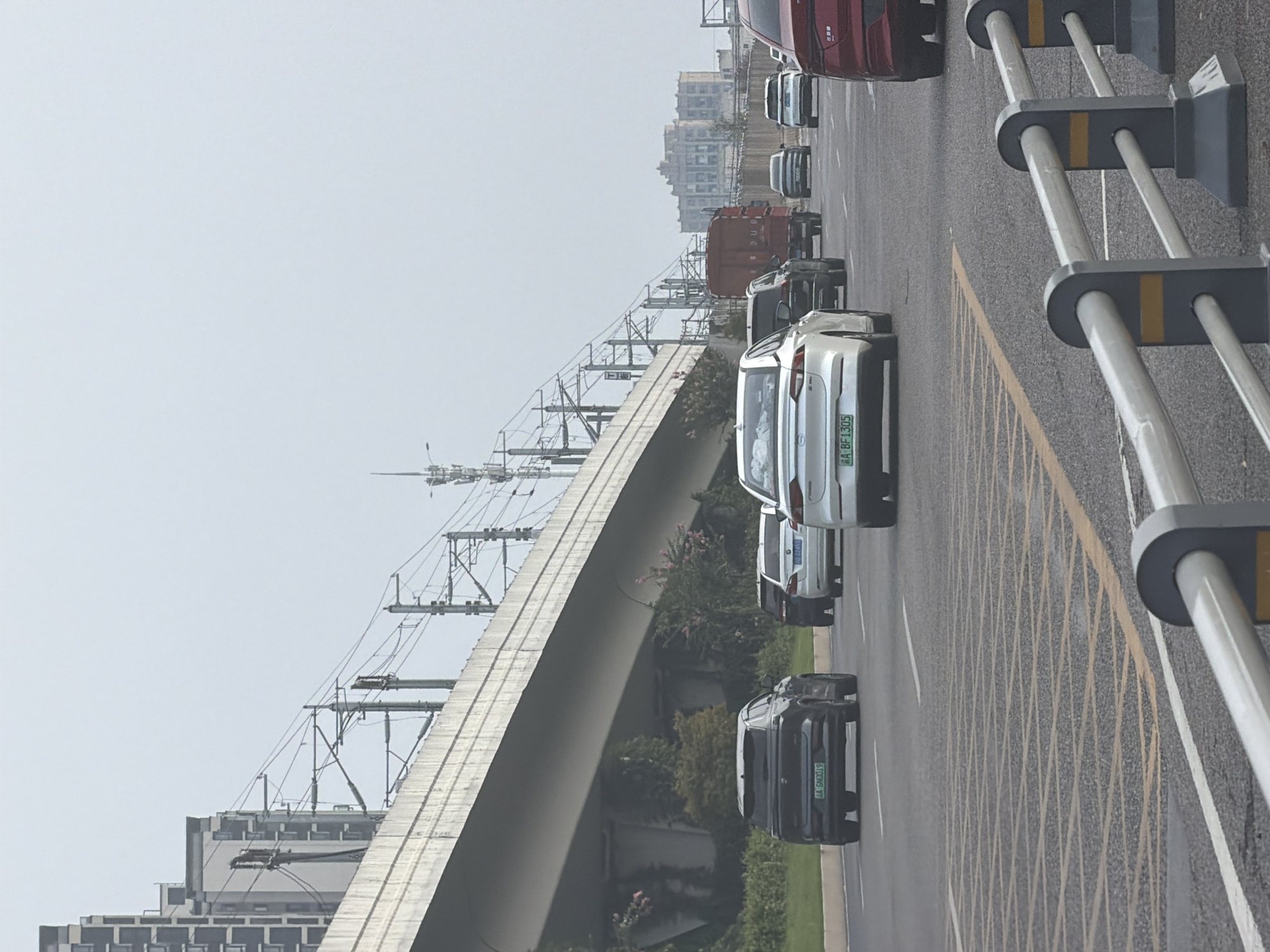 The image captures a street scene in Jiangjin, China, with vehicles traveling on a multi-lane road. In the foreground, a road barrier with white and yellow markings is visible. Several cars are present, including a black SUV with license plate "AA GN0019", a white sedan with license plate "A BF1305", and a red car further down the road. Other vehicles, including a red truck, are also in motion. An elevated road or bridge runs overhead, supported by concrete structures and flanked by overgrown vegetation. Electrical infrastructure, including overhead wires and poles, is visible along the elevated structure, suggesting the presence of a railway or tram line. In the background, tall buildings and smaller structures can be seen, indicating an urban environment. The sky is overcast, suggesting a cloudy day.
