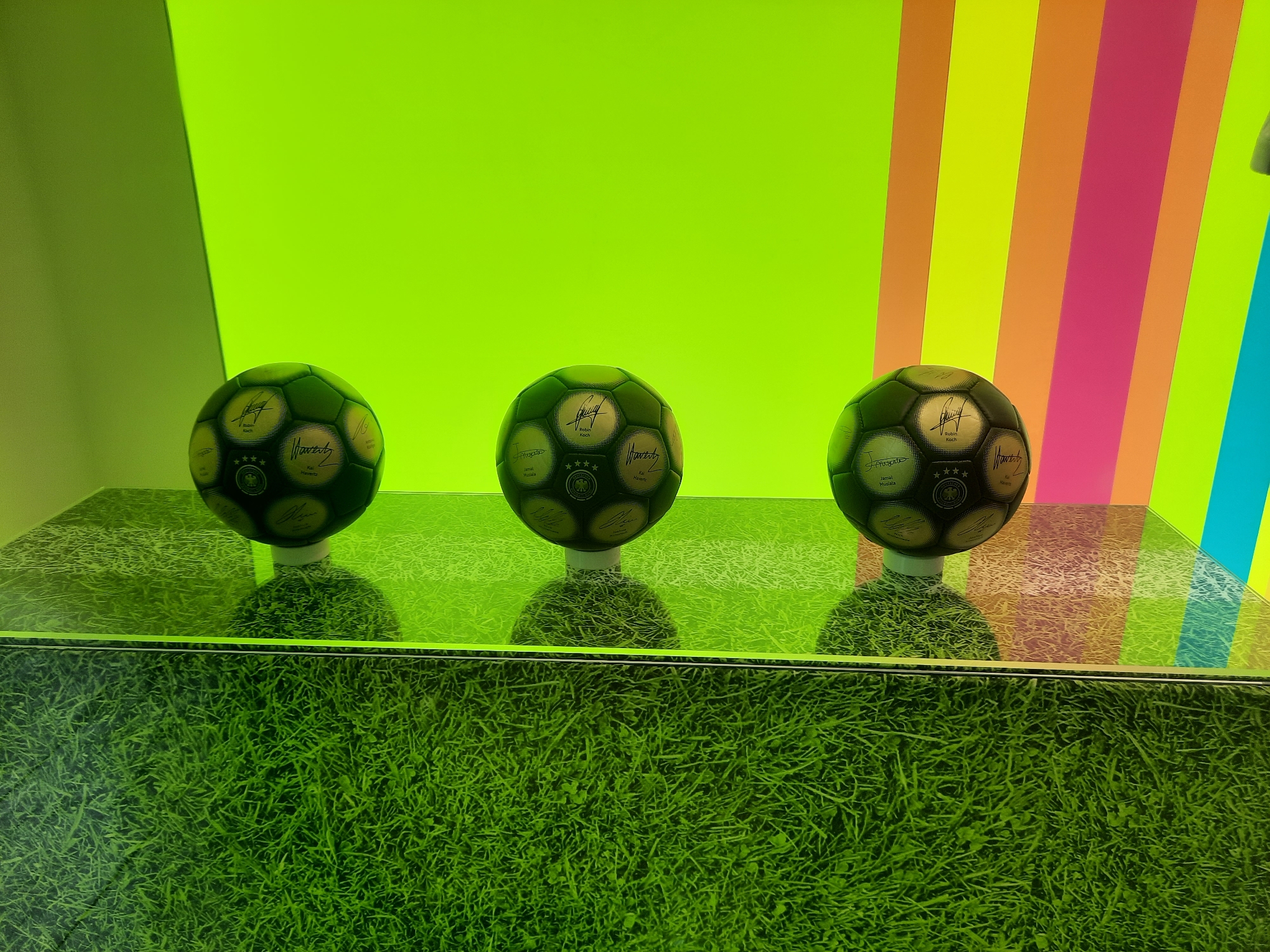 The image displays three autographed soccer balls arranged in a row on a reflective surface resembling grass. The background consists of two distinct sections: the left side is a solid bright lime green, while the right side features a vertical striped pattern in various bright colors including yellow, orange, pink, and blue. Each soccer ball is black and gold, with signatures and text printed on its panels. The balls are positioned on a display case which has a grass-like texture on its front. The overall lighting is bright and vibrant. The context of this display is at the "Fussballmuseum" (Football Museum) in Rosenheim, Germany, where these autographed balls are part of an exhibition.