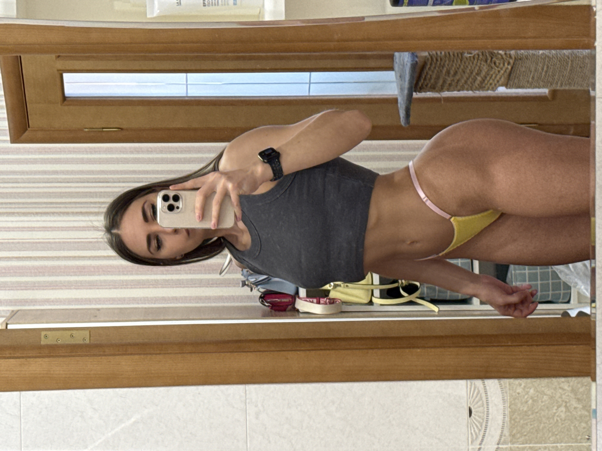 The image, rotated 90 degrees clockwise, displays a young woman taking a mirror selfie indoors. She has long dark hair and is wearing a dark gray cropped tank top and yellow thong bikini bottoms with thin pink straps. On her left wrist, she sports a dark smartwatch. She holds a light-colored smartphone, likely an iPhone, in her right hand, capturing her reflection with a neutral expression.
The background, reflected in the mirror, shows an indoor setting. Above the subject, a wooden structure resembling a door or cabinet with a window pane is visible. Behind her, various items are reflected, including a textured mat or rug, and a few bags, one yellow and another pink/red. The wall in the reflection features striped wallpaper, and the floor below the mirror is tiled, displaying some decorative patterns. Faint, blurry text is visible on some reflected objects in the background, such as "Essi" and "till" on what appears to be packaging or labels. The scene suggests a private space, possibly a bathroom or dressing area. No specific visual elements within the image directly indicate the provided location context of Saint Petersburg, Russia.