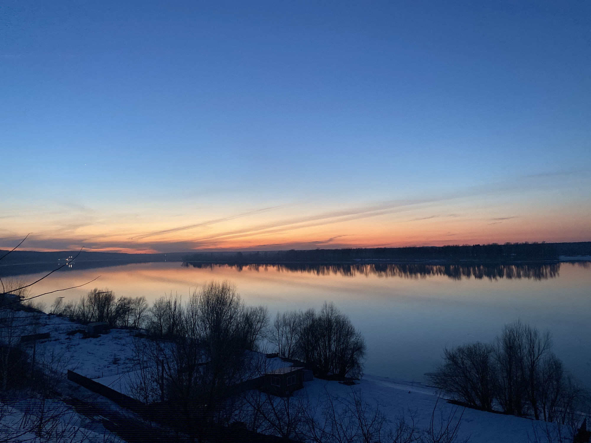 The image shows a serene landscape at twilight. The sky transitions from a deep blue at the top to a soft orange glow near the horizon. Wispy clouds stretch across the sky. Below, a wide body of water reflects the colors of the sky, creating a symmetrical effect. The opposite shore is lined with trees, their dark silhouettes mirrored in the water. In the foreground, the ground is covered in snow. Bare trees and bushes dot the landscape, and a small building is partially visible amidst the snow and foliage. The location is possibly near the city of Ольховка, Russia, based on the environment.