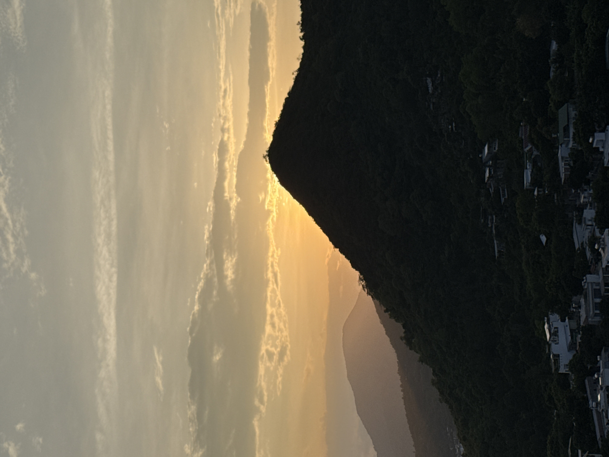 This is a photograph of a mountainous landscape at sunset. The sun is partially obscured by a silhouetted mountain peak, casting a warm, golden glow across the sky. The sky is filled with wispy clouds, some of which are illuminated by the setting sun. In the foreground, the dark silhouette of a densely forested mountain slope is visible, with scattered houses and buildings nestled among the trees. The overall impression is one of natural beauty and tranquility, with a hint of human habitation blending into the landscape. The location is identified as Bac Nha Trang Ward, Vietnam.